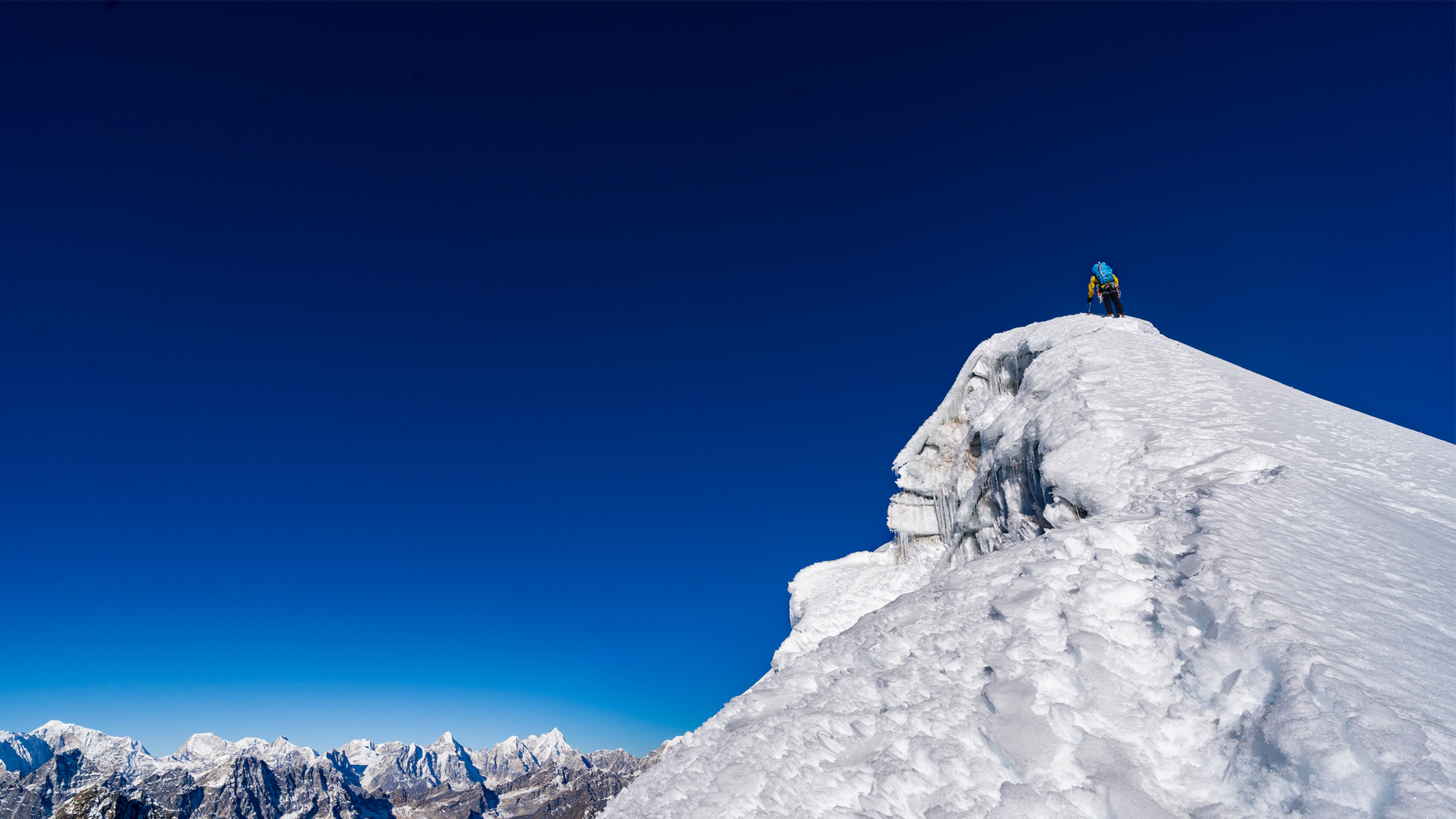 Climber ascending a snowy mountain peak under a clear blue sky, with distant mountain ranges visible in the background.