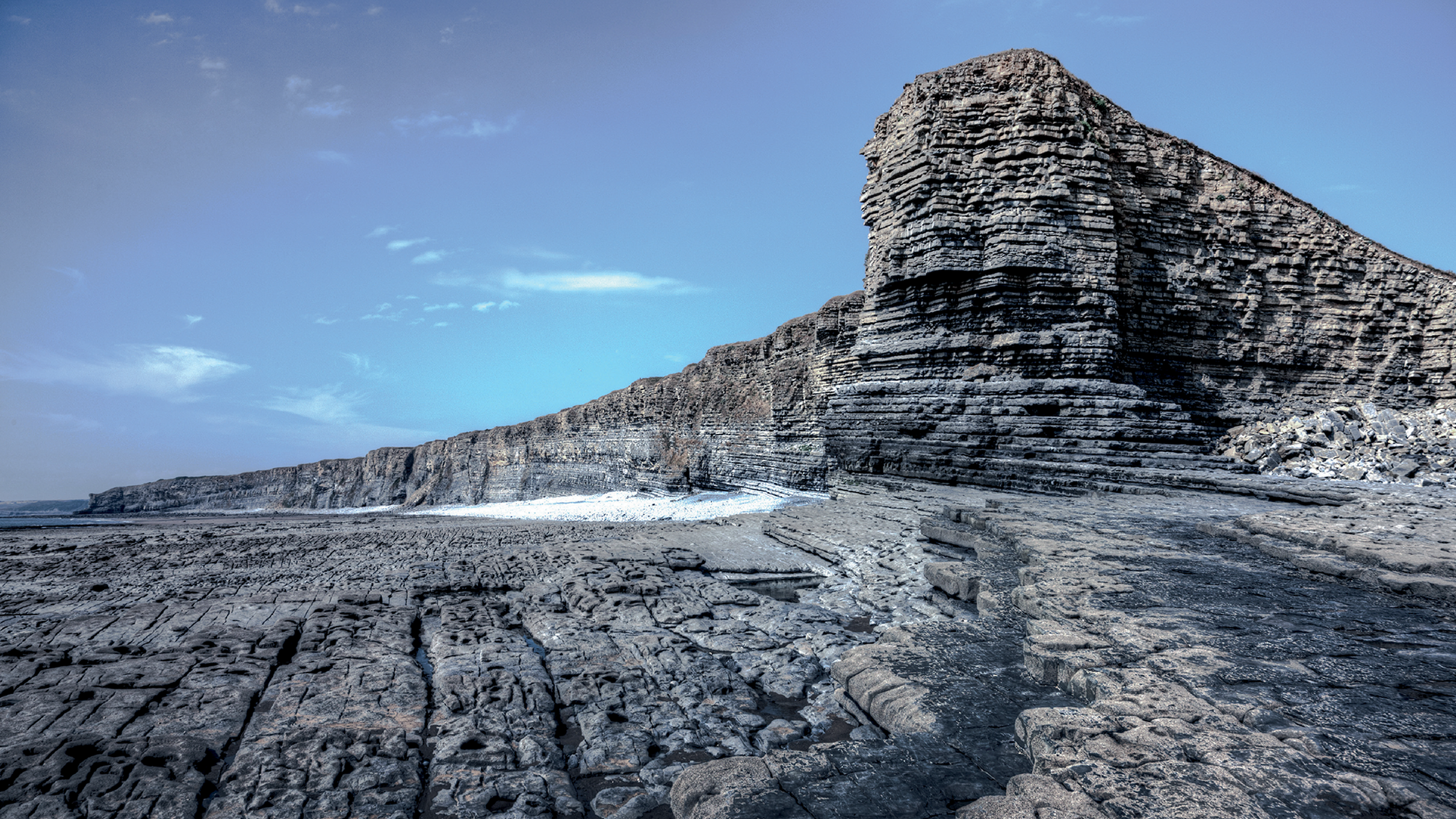 Rocky cliff face with layered formations under a clear sky, extending along a rugged coastal landscape.