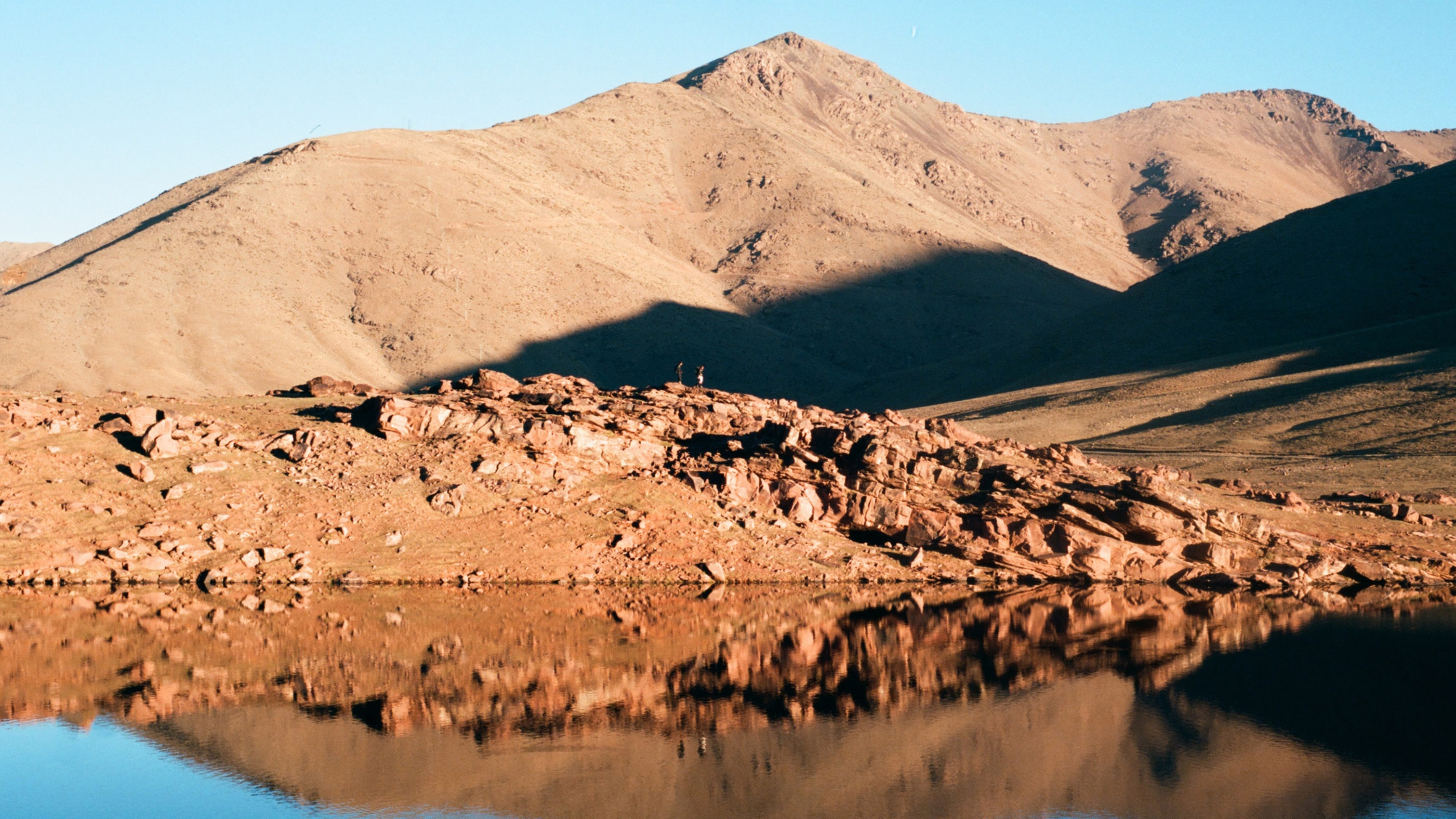 Two people stand on rocky terrain by a calm lake, with arid mountains in the background under a clear blue sky.