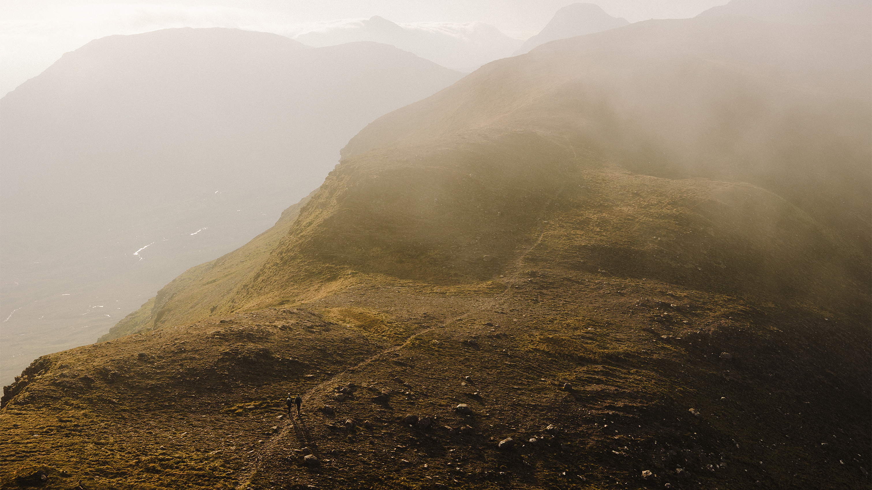 Misty mountain ridge with two hikers walking on a narrow trail. Low visibility creates an atmospheric, serene landscape.