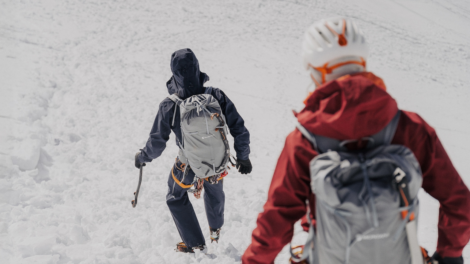 Two climbers in winter gear and helmets trek through snow, carrying ice axes and wearing backpacks, with a snowy landscape in the background.