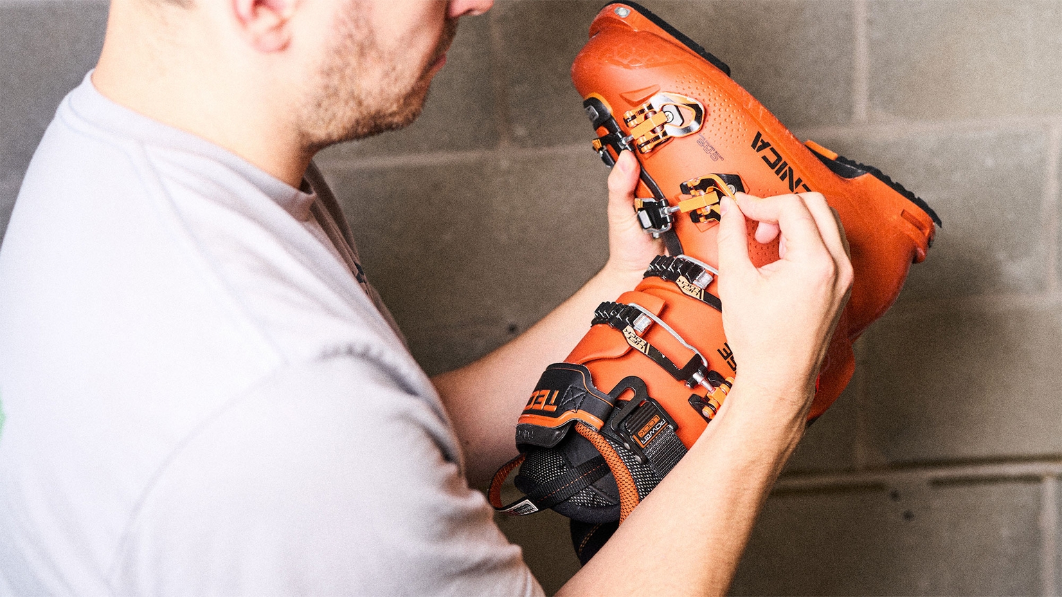 Person adjusting buckles on an orange ski boot, wearing a light gray shirt, against a concrete wall background.