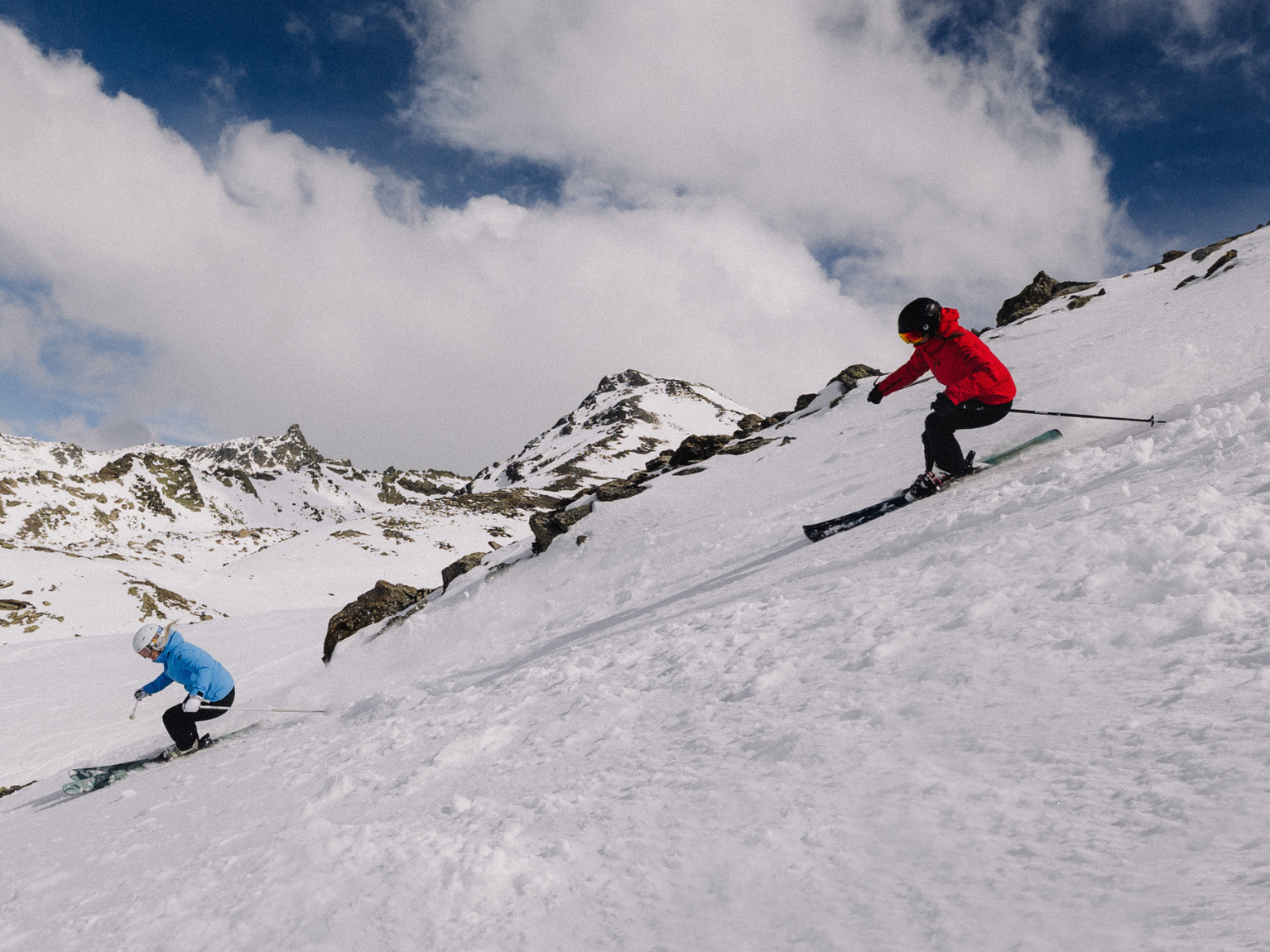 Two skiers in blue and red jackets descend a snowy mountain slope under a partly cloudy sky, surrounded by rugged peaks.