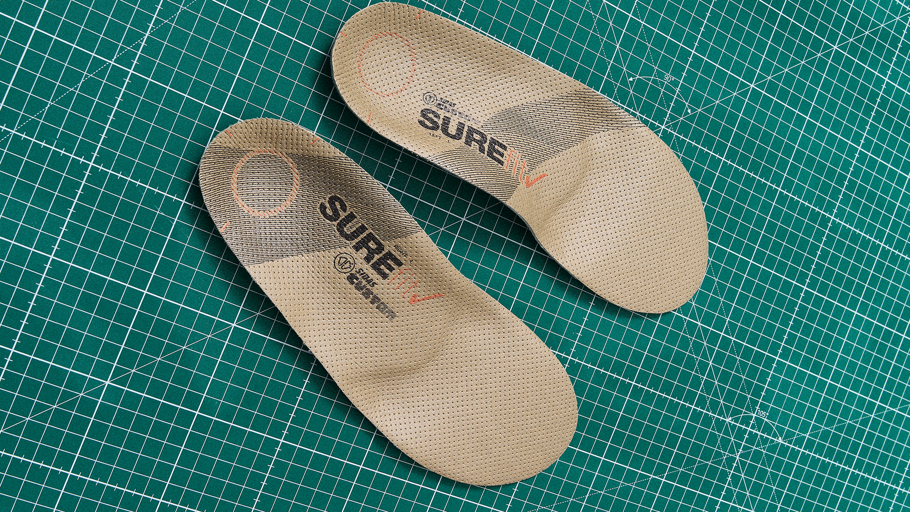 Two beige insoles with branding on a green grid cutting mat.