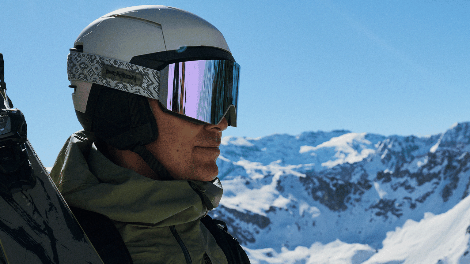 A skier wearing a helmet and reflective goggles stands against a backdrop of snow-covered mountains under a clear blue sky.