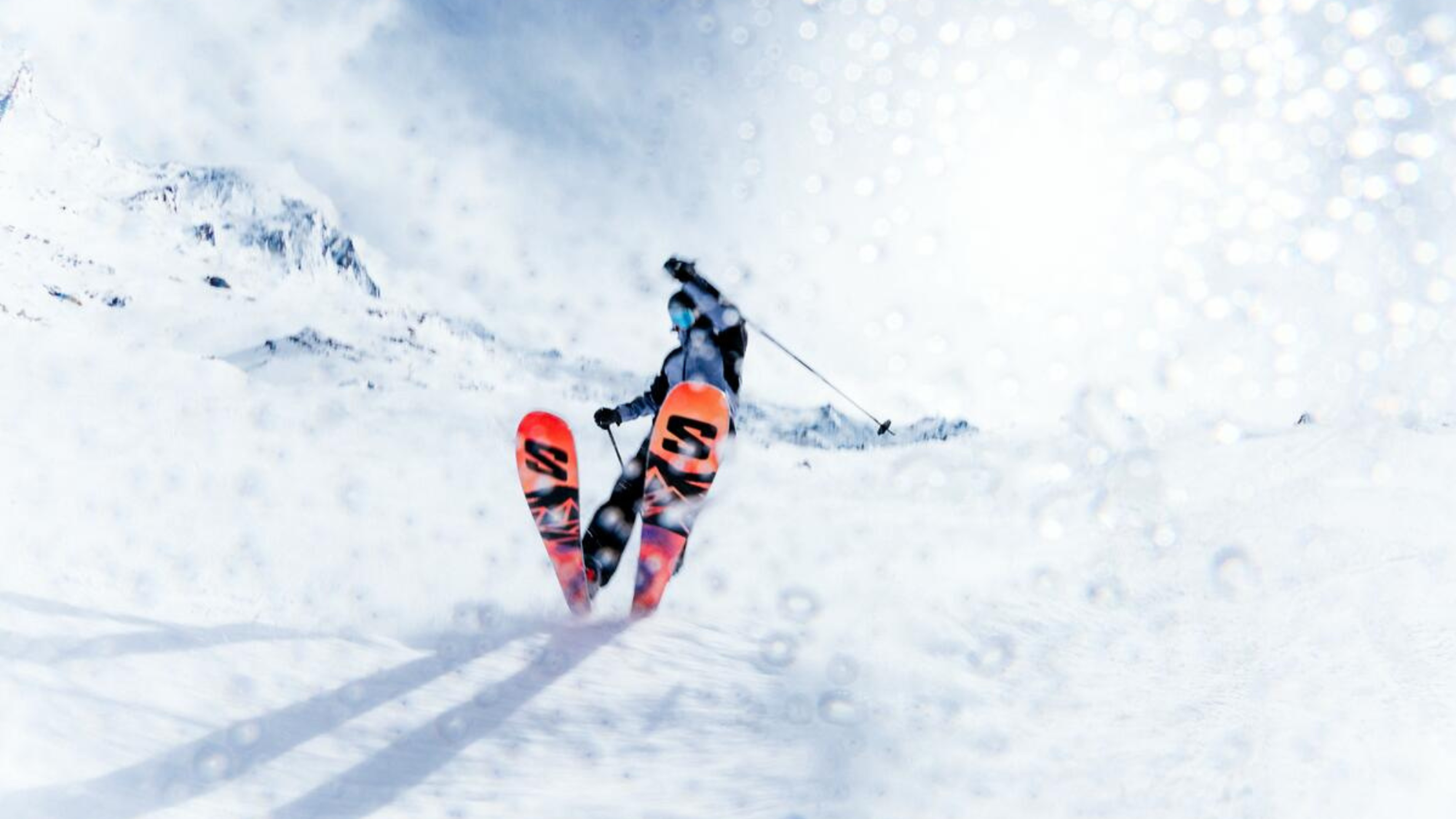 Skier in mid-air performing a jump on snowy mountain terrain, with bright orange skis against a backdrop of clear blue sky.