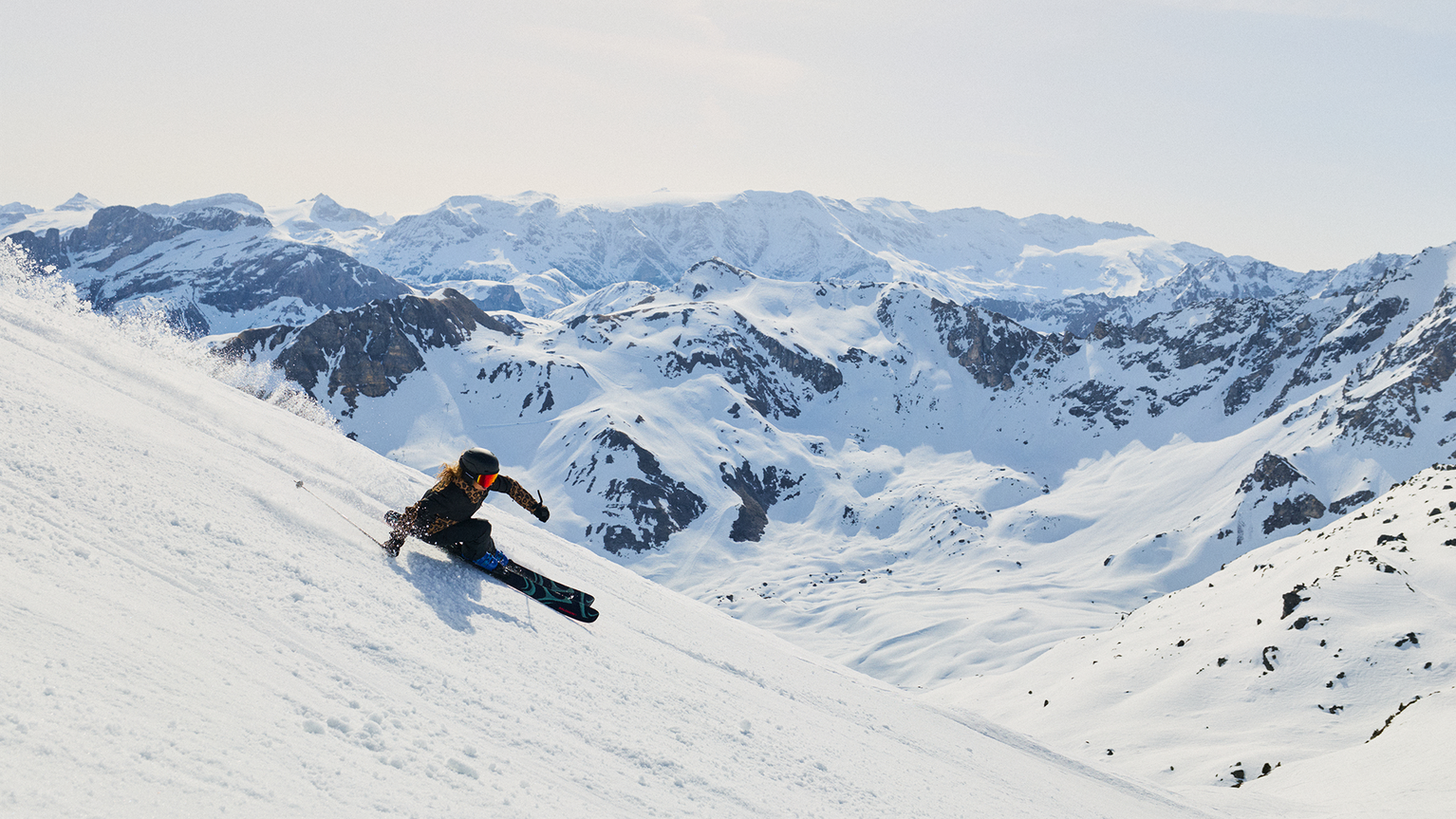 A skier expertly navigates a steep, snowy slope with stunning, rugged mountain peaks in the background under a clear sky.