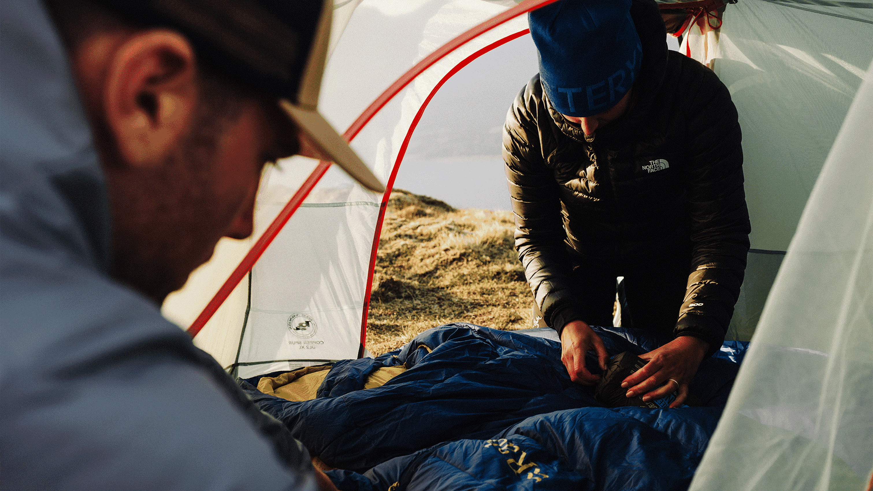 Two people setting up a sleeping bag inside a camping tent on a grassy hillside.