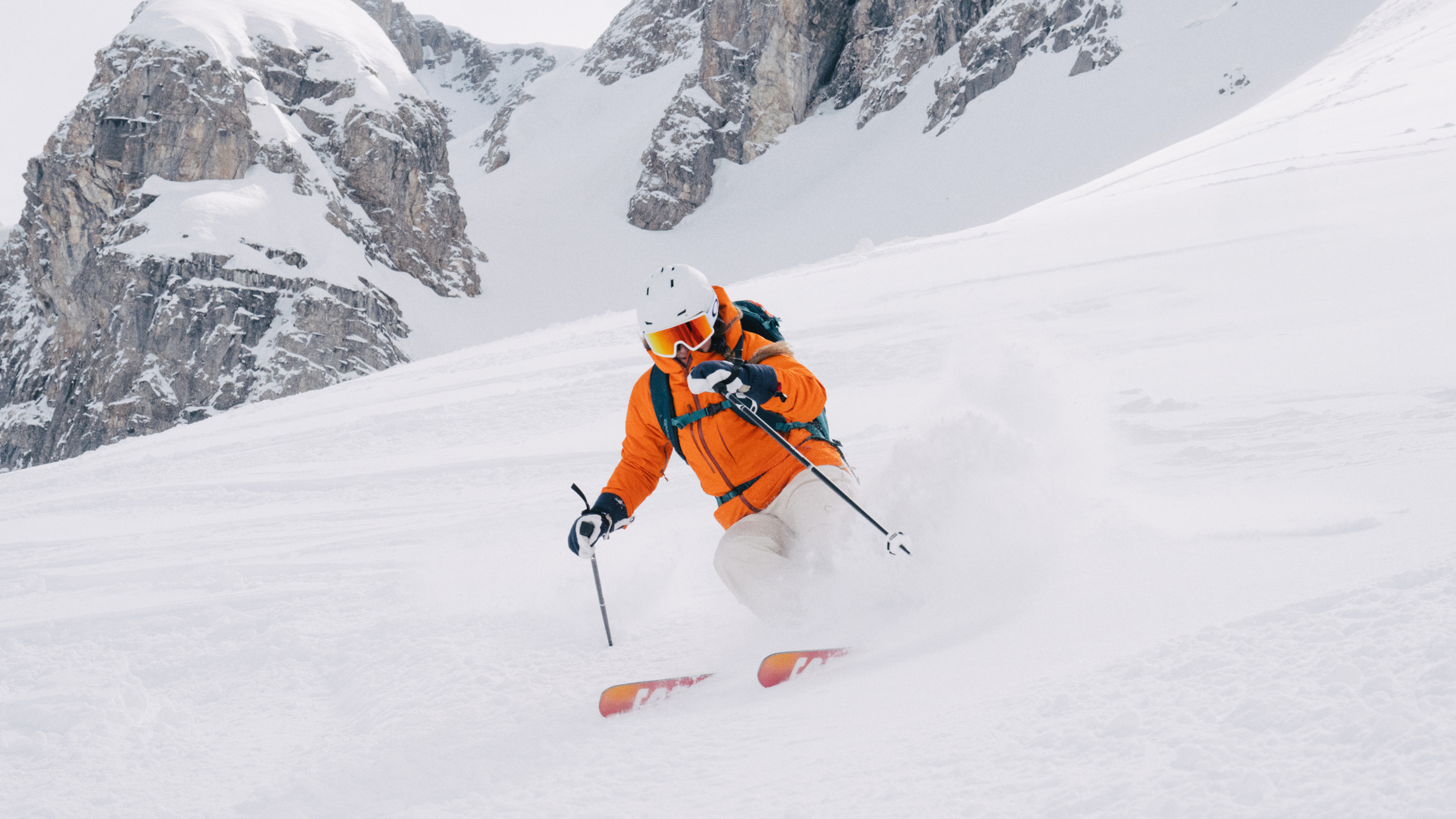 Skier in an orange jacket and helmet navigating through fresh snow on a mountainous slope.