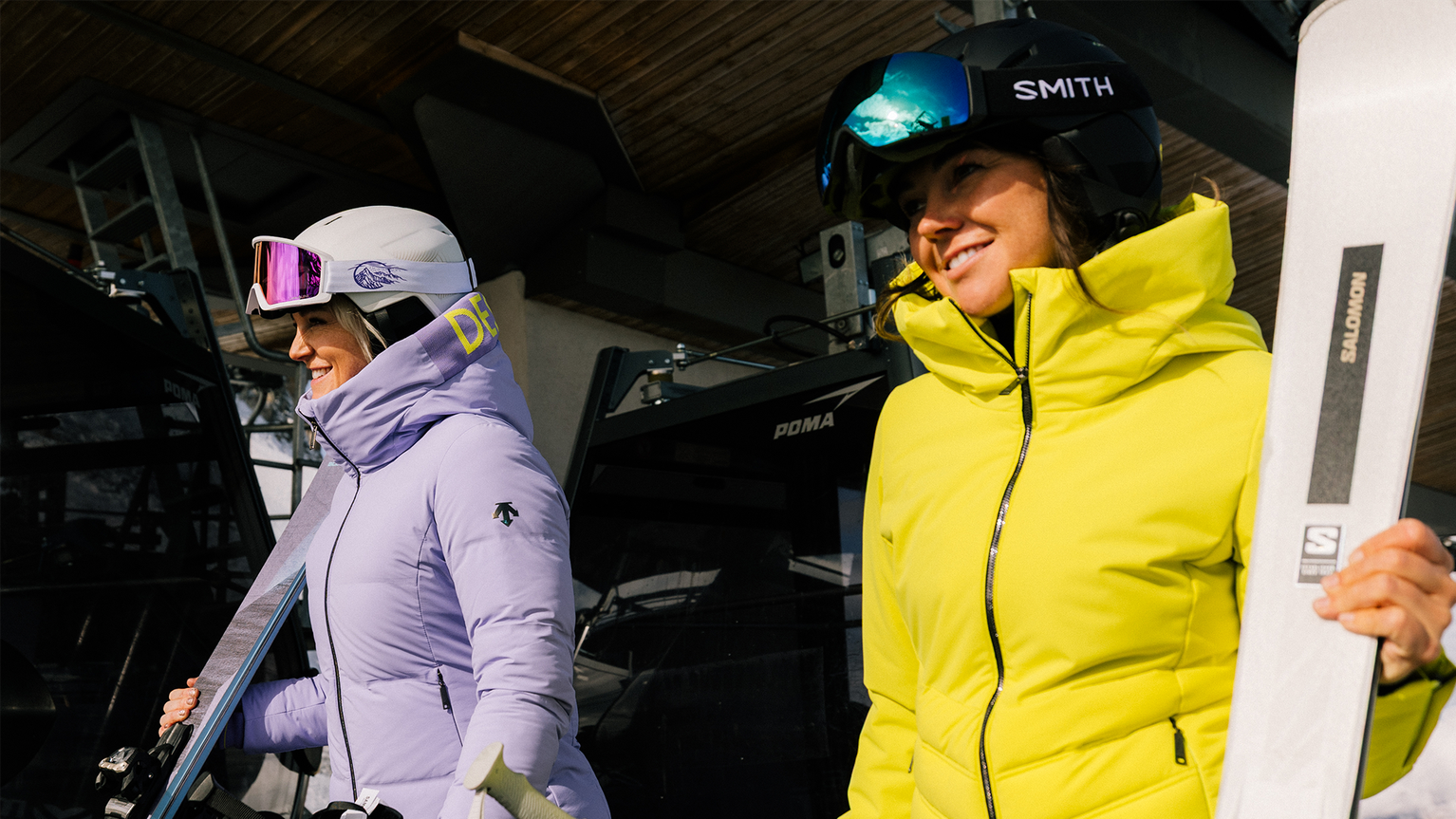 Two skiers in pastel purple and neon yellow jackets carry skis and poles, smiling beside a chairlift.