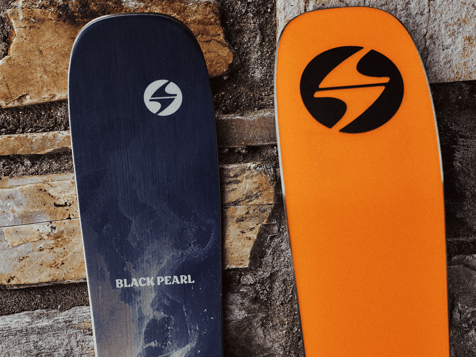 Two skis leaning against a stone wall, one blue marked "Black Pearl" and one orange, both featuring a circular logo.