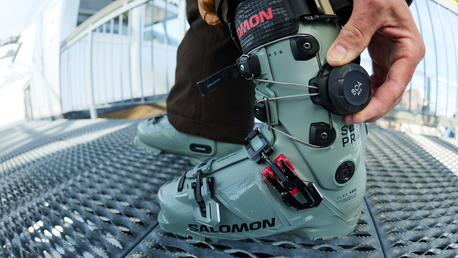 Adjusting grey ski boots with black and red buckles on a metal grated surface, showcasing precision and quality for mountain sports enthusiasts.