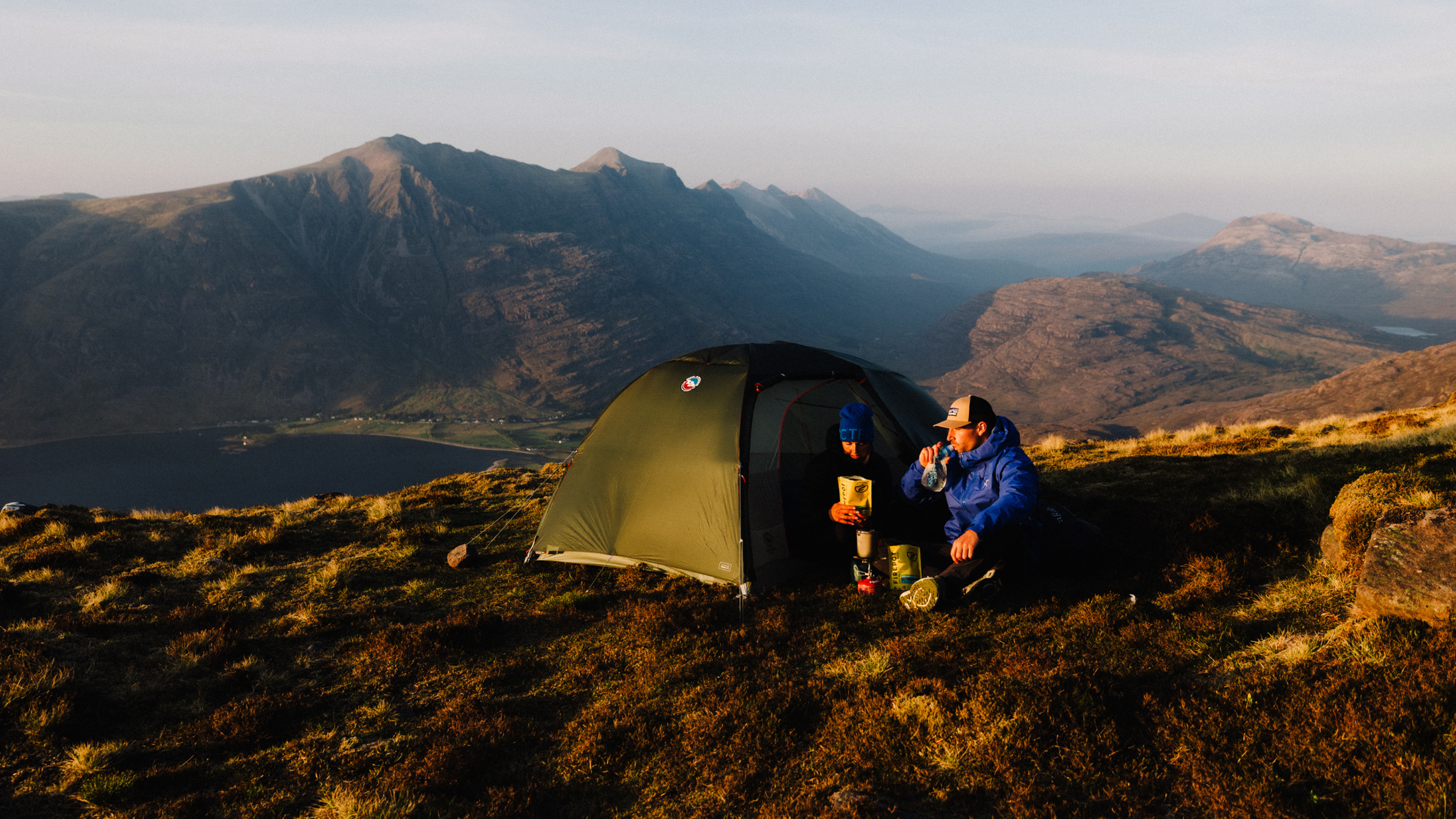 Two campers sit by a green tent on a grassy mountain, enjoying a drink, with expansive mountain views in the background at sunset.
