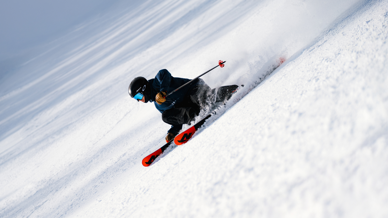 Skier in dark gear carving through fresh snow on a slope, with red skis and poles, against a bright, snowy backdrop.