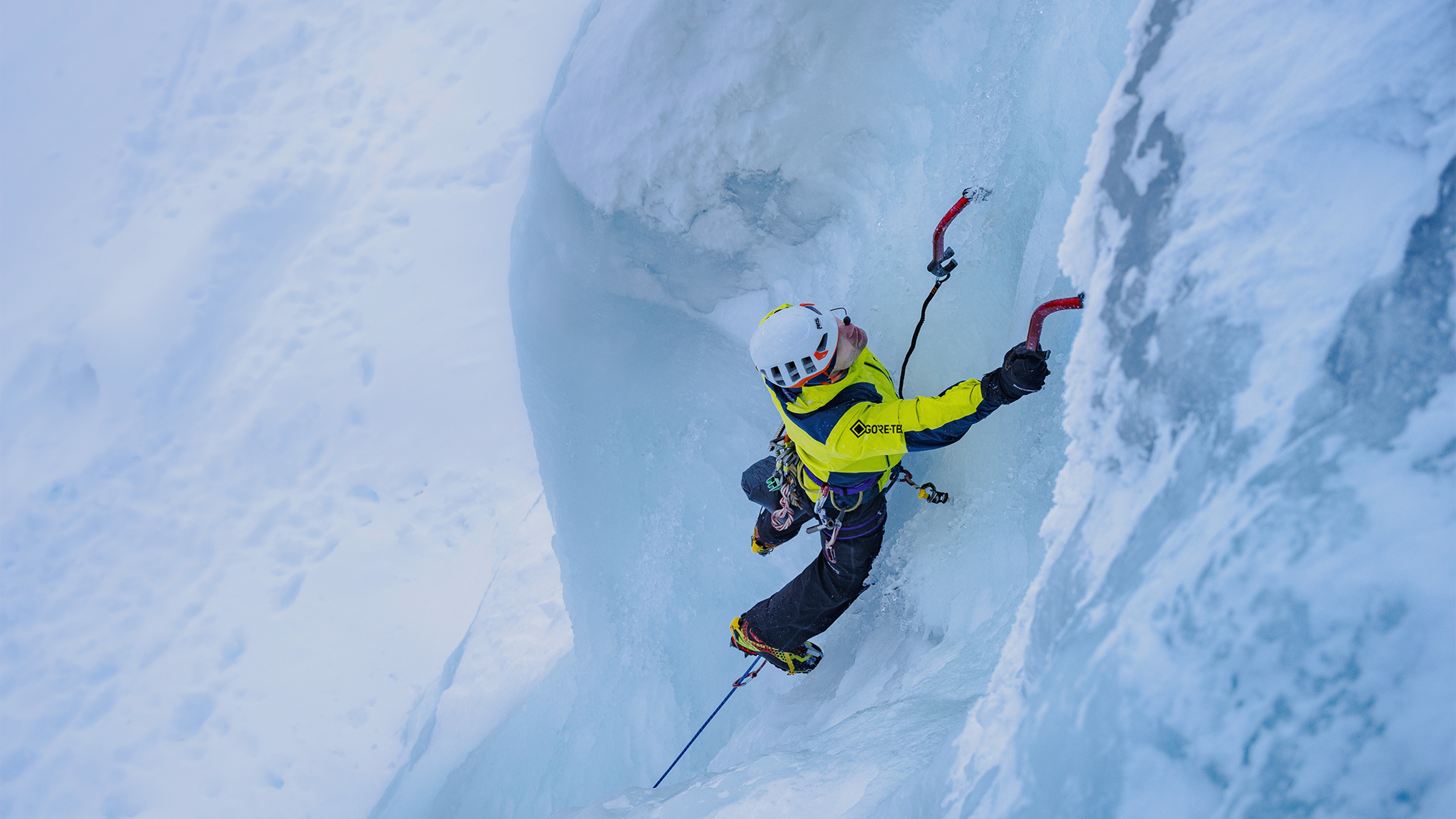 Ice climber in bright yellow jacket and white helmet using ice axes and crampons on a steep blue ice wall.