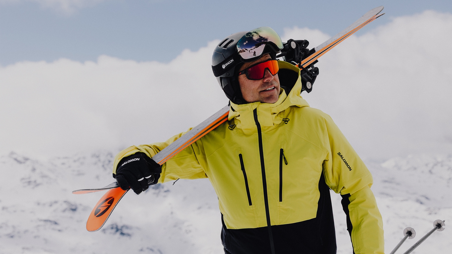 Man in yellow ski jacket and helmet carrying skis on shoulder, standing on snowy mountain with cloudy sky backdrop.
