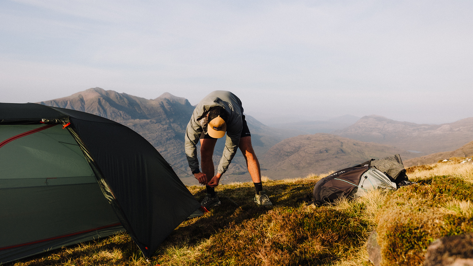Camper setting up a tent on a grassy mountain ridge with a scenic view of rolling hills in the background.