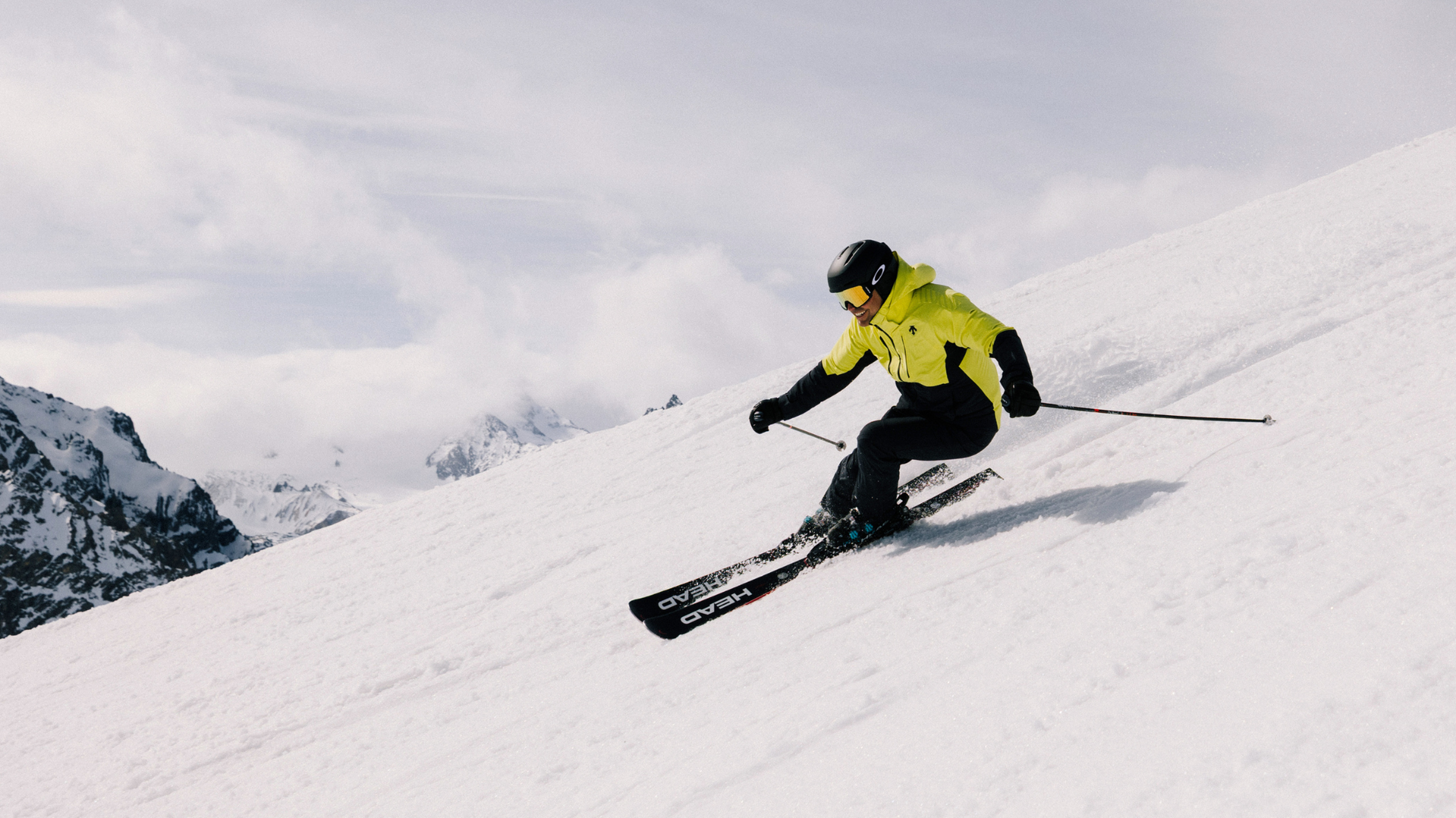 Person skiing down a snowy mountain slope, wearing a bright yellow jacket and black helmet, with snow-covered peaks in the background.