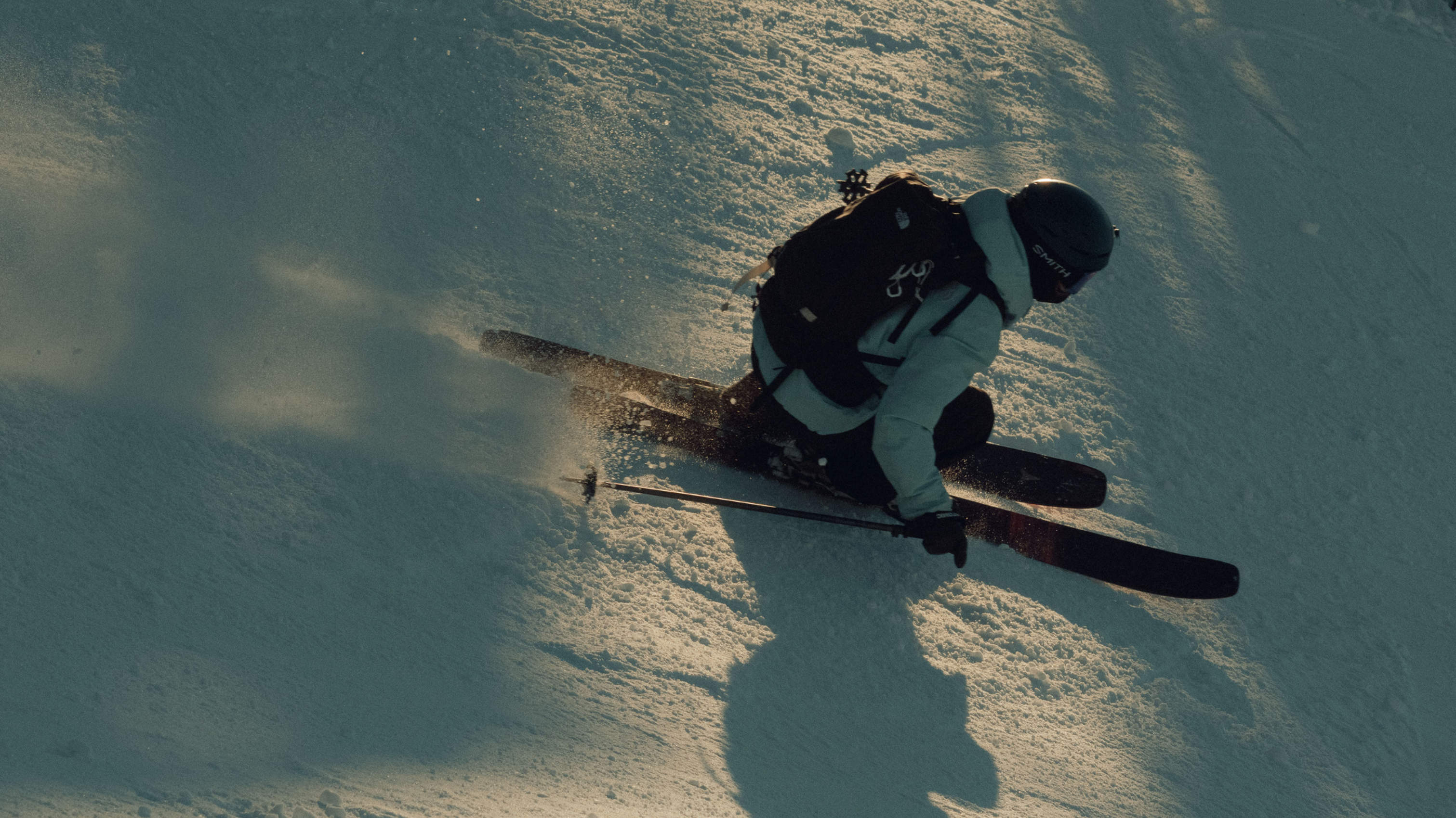 Skier in gear gliding down a snowy slope with a backpack, ski poles in hand, casting a shadow on the snow.
