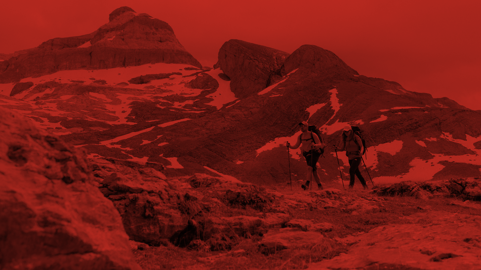Two hikers traverse a rocky mountain landscape, using poles, under a red-tinted sky. Snow patches are visible on the rugged terrain.