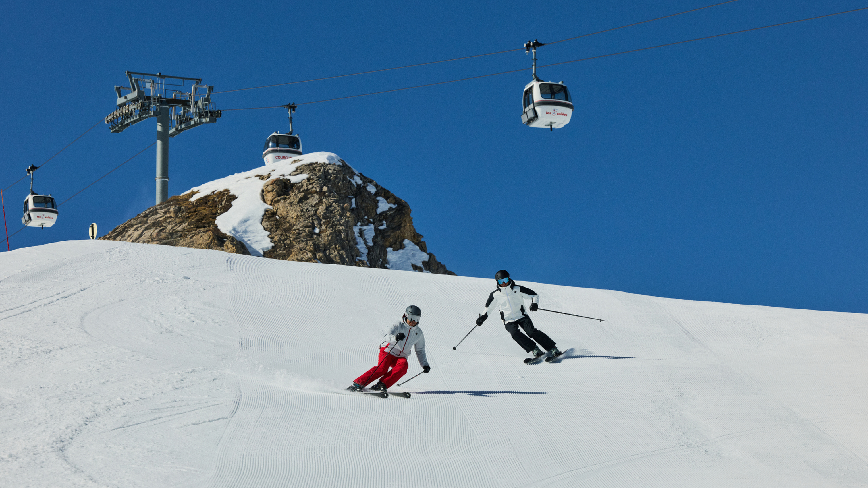 Two skiers navigate a snowy slope under blue skies and cable cars in the background.