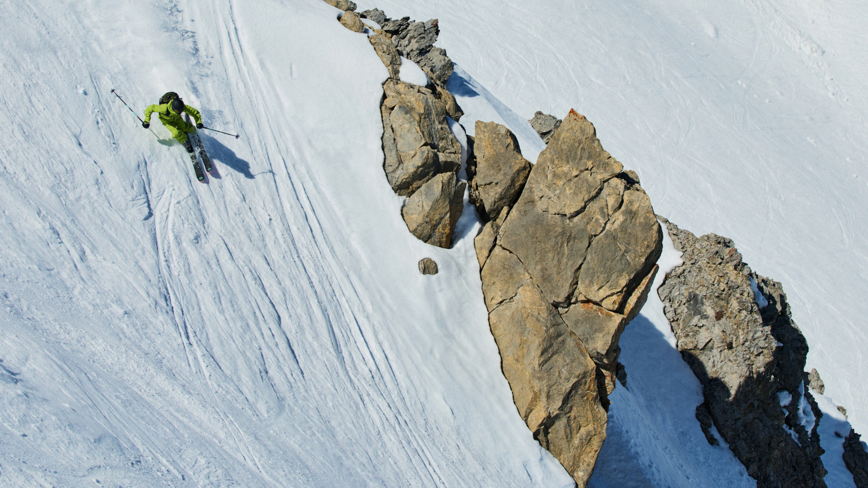 Skier in green Norrona ski gear descends a steep snowy slope beside jagged rocks.