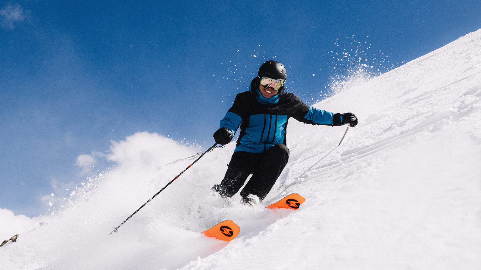 Person skiing downhill on a snowy slope, wearing a blue and black jacket, helmet, and goggles, with bright orange skis.