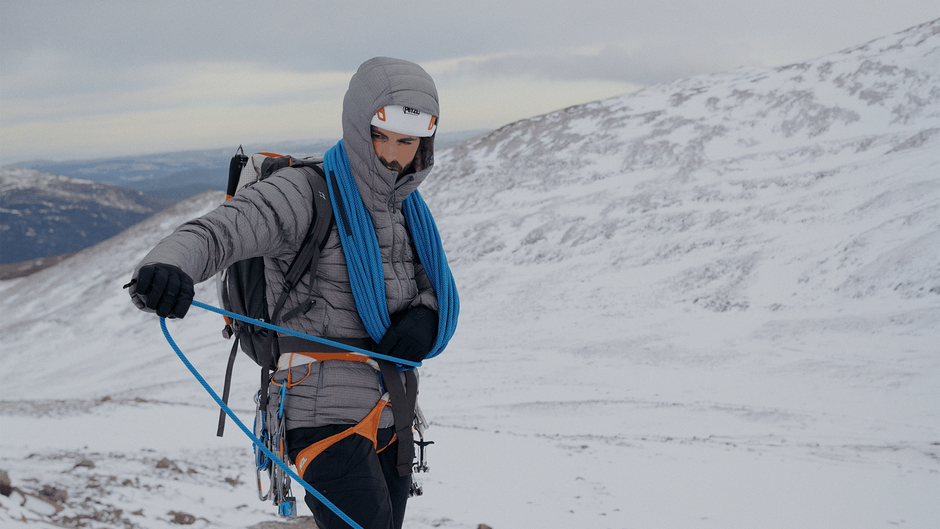 An individual dressed in mountaineering gear, holding a blue climbing rope, stands on a snowy mountain landscape.