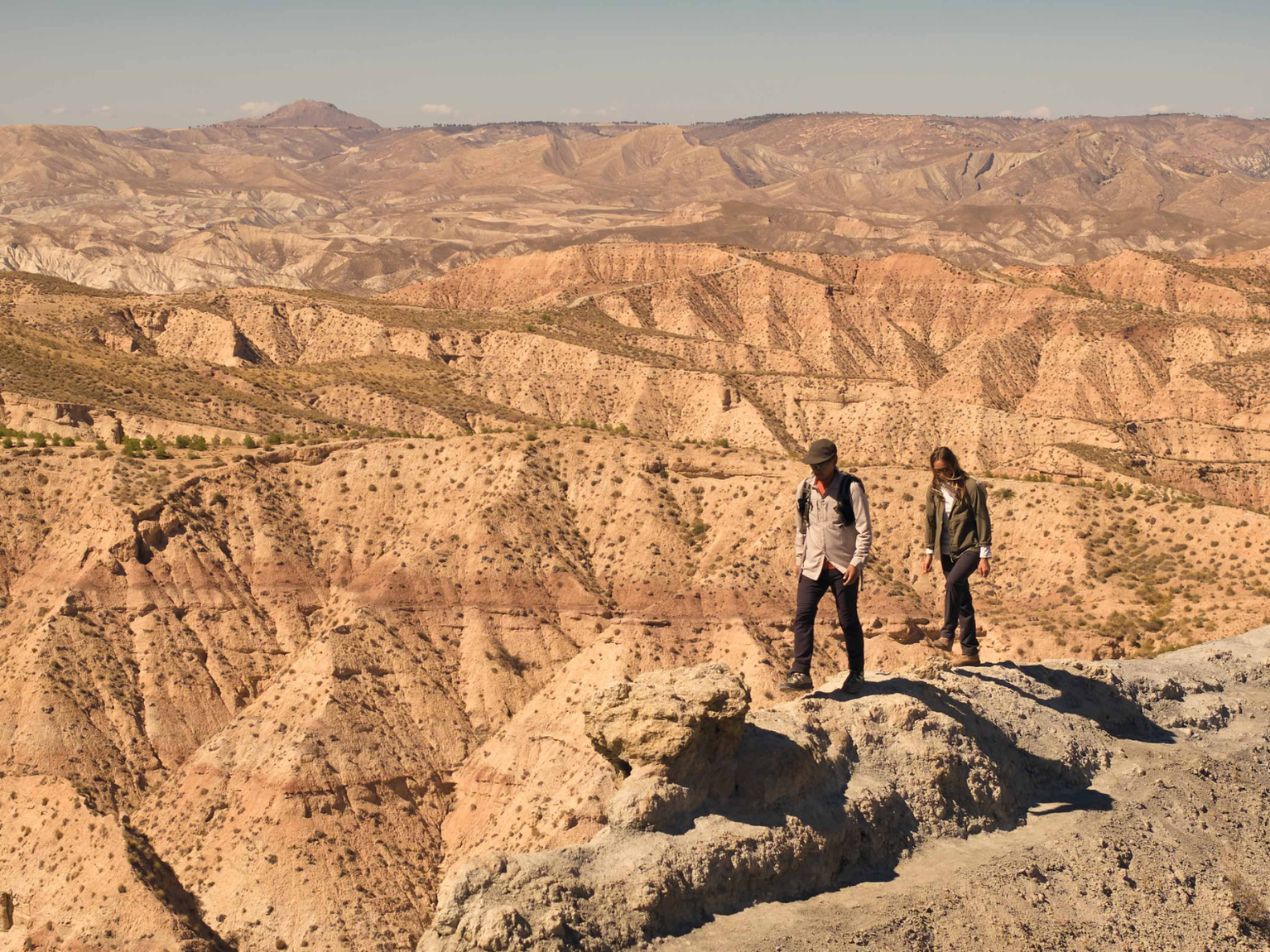 Two people stand on a rocky cliff, overlooking a vast, rugged desert landscape under a clear sky.