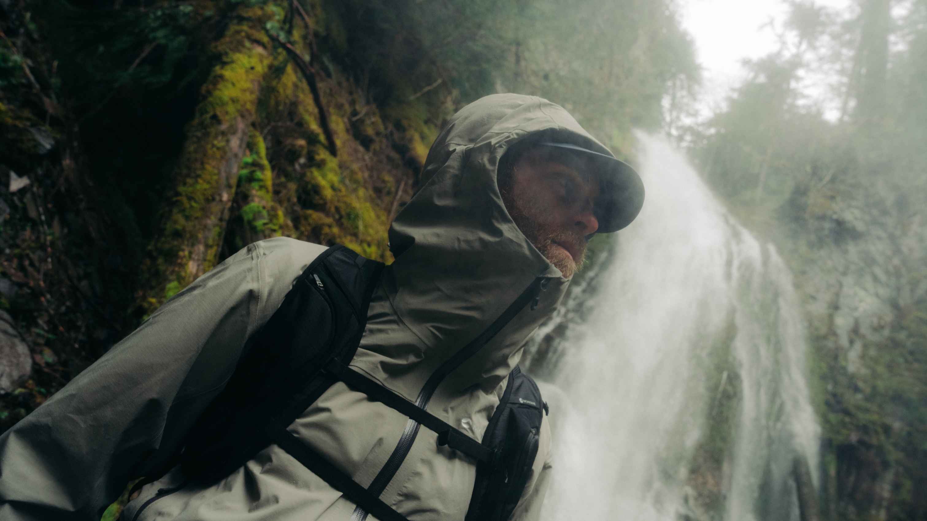 Man in weatherproof jacket hiking near a waterfall in a lush, misty forest.