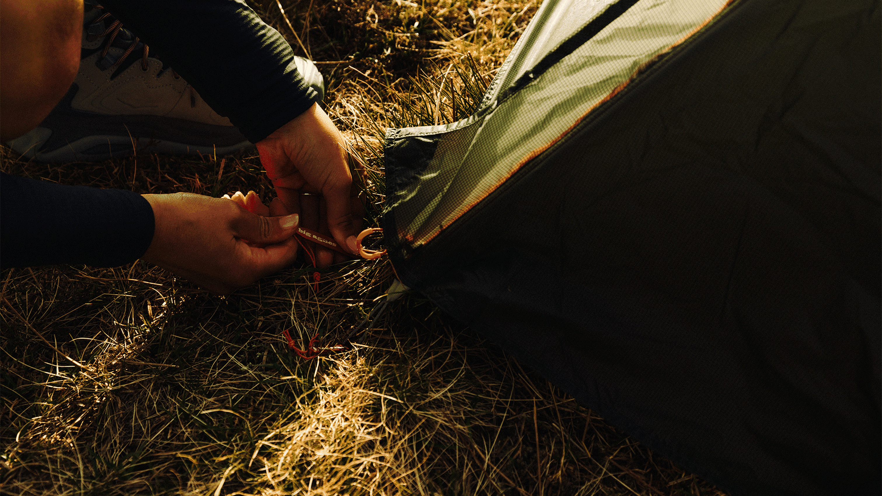 Hands fastening a tent to the ground with stakes on grassy terrain, wearing hiking boots.