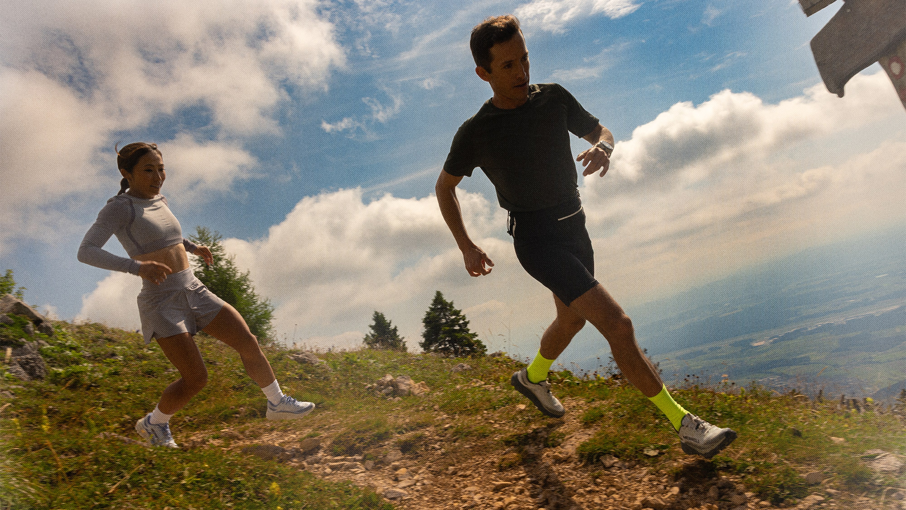 Two people running on a mountain trail, wearing athletic clothing. Bright sky and expansive landscape in the background.