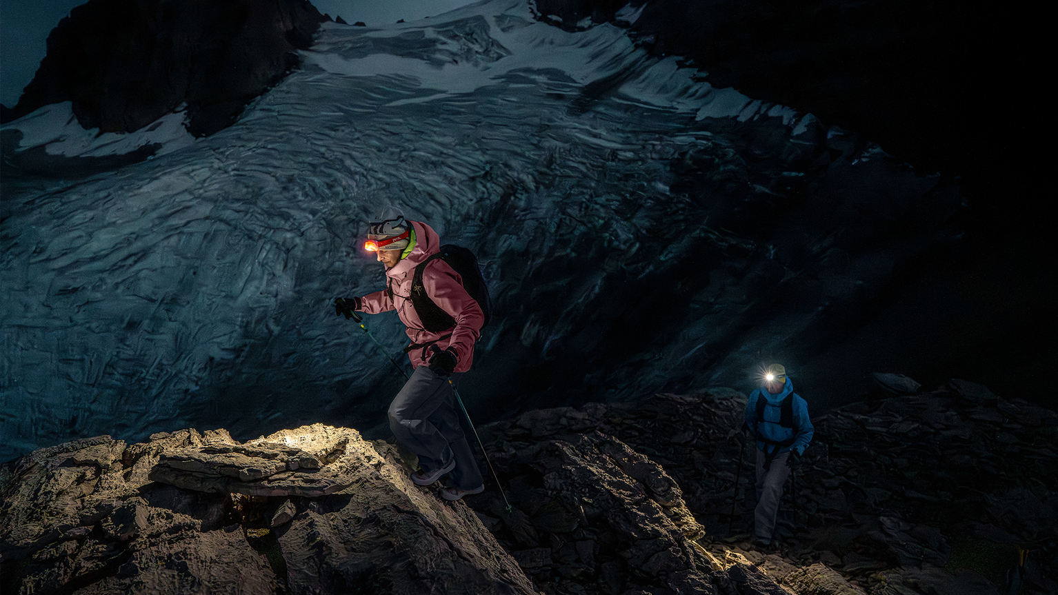 Two climbers with headlamps navigate a rocky mountain path at night, snowy peaks visible in the background, showcasing adventure and exploration.