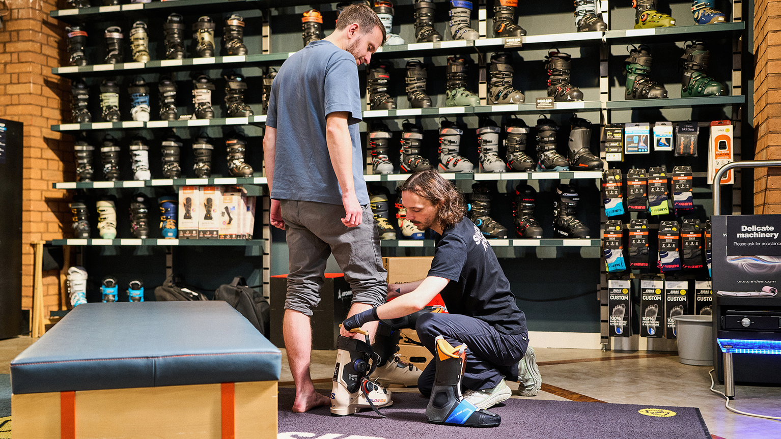 A person is being fitted for ski boots by a store employee in a shop with shelves full of various ski boots in the background.