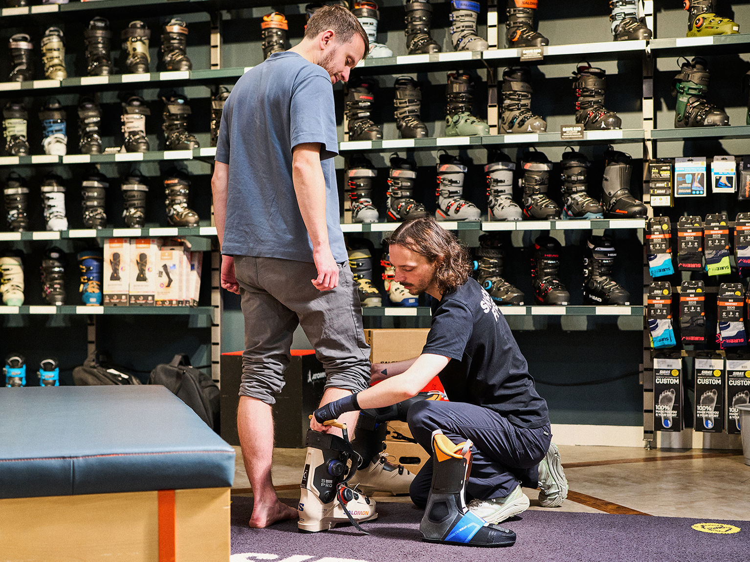 A person is being fitted for ski boots by a store employee in a shop with shelves full of various ski boots in the background.