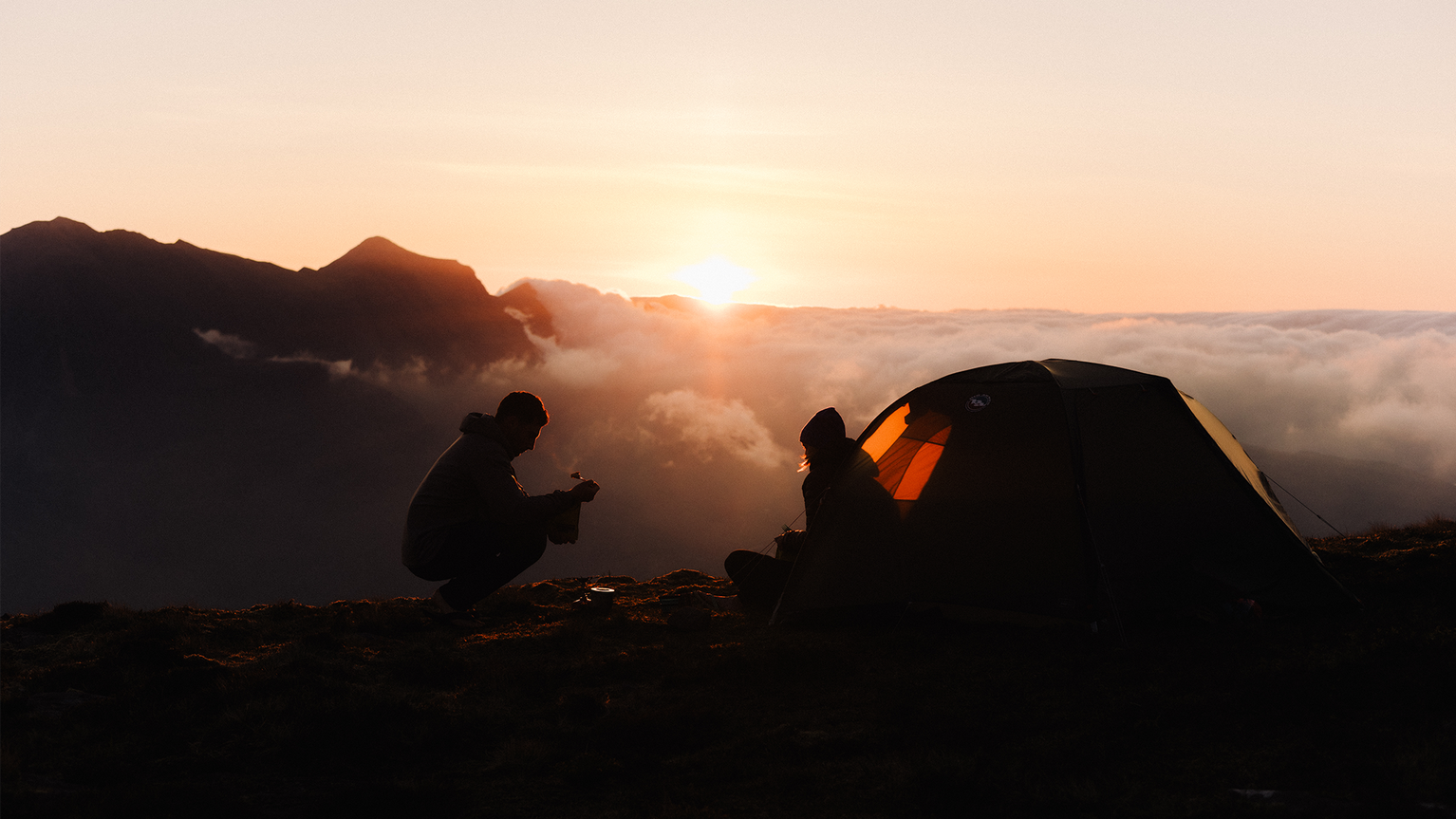 Silhouettes of two people and a tent on a mountain peak at sunset, with clouds and distant mountains in the background.