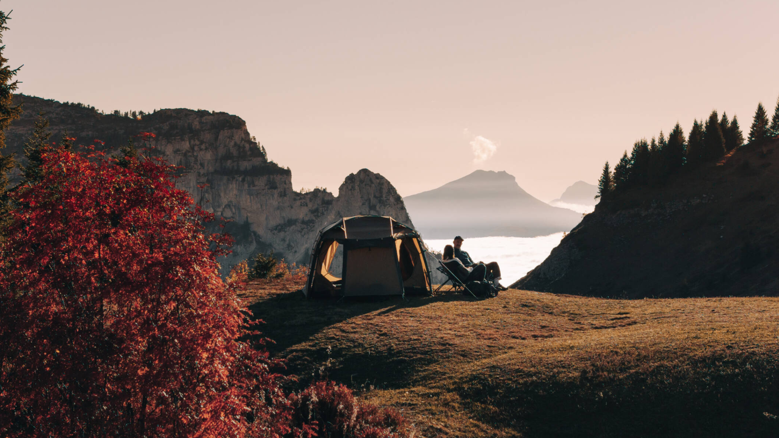 Two campers relax by a tent on a mountain ridge, with a breathtaking view of distant peaks and a valley blanketed in clouds.