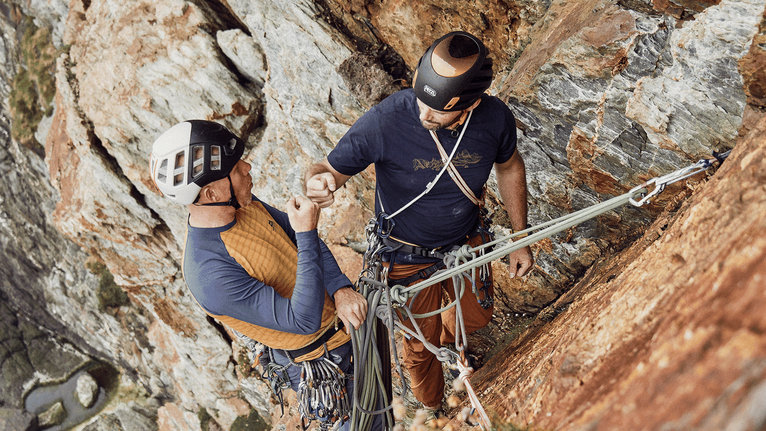 Two climbers sharing a fist bump on a rugged rock face, wearing helmets and harnesses, with ropes securely attached.