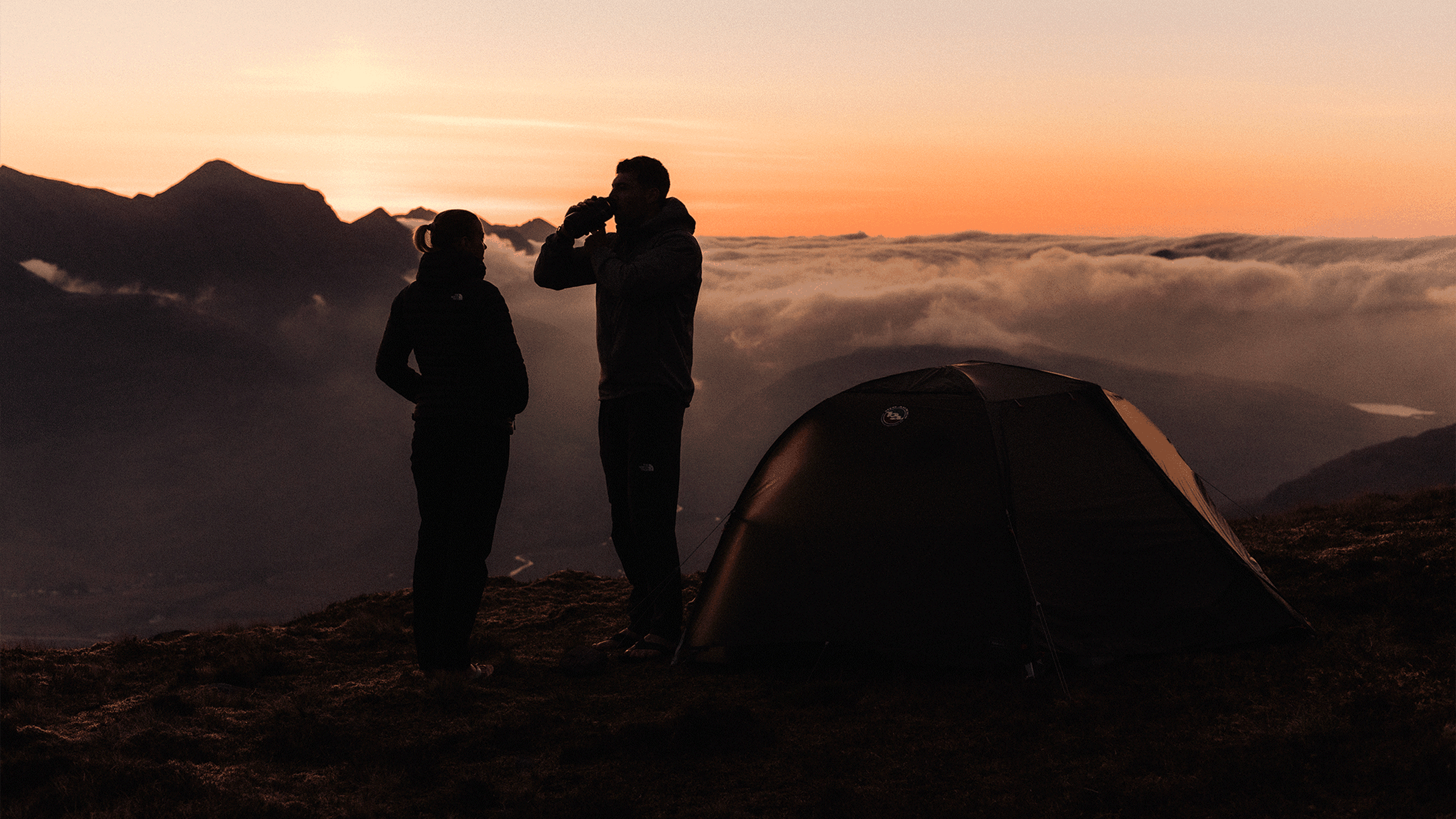 Two campers stand near a tent at sunrise, surrounded by mountains and clouds. One drinks from a cup, enjoying the view.