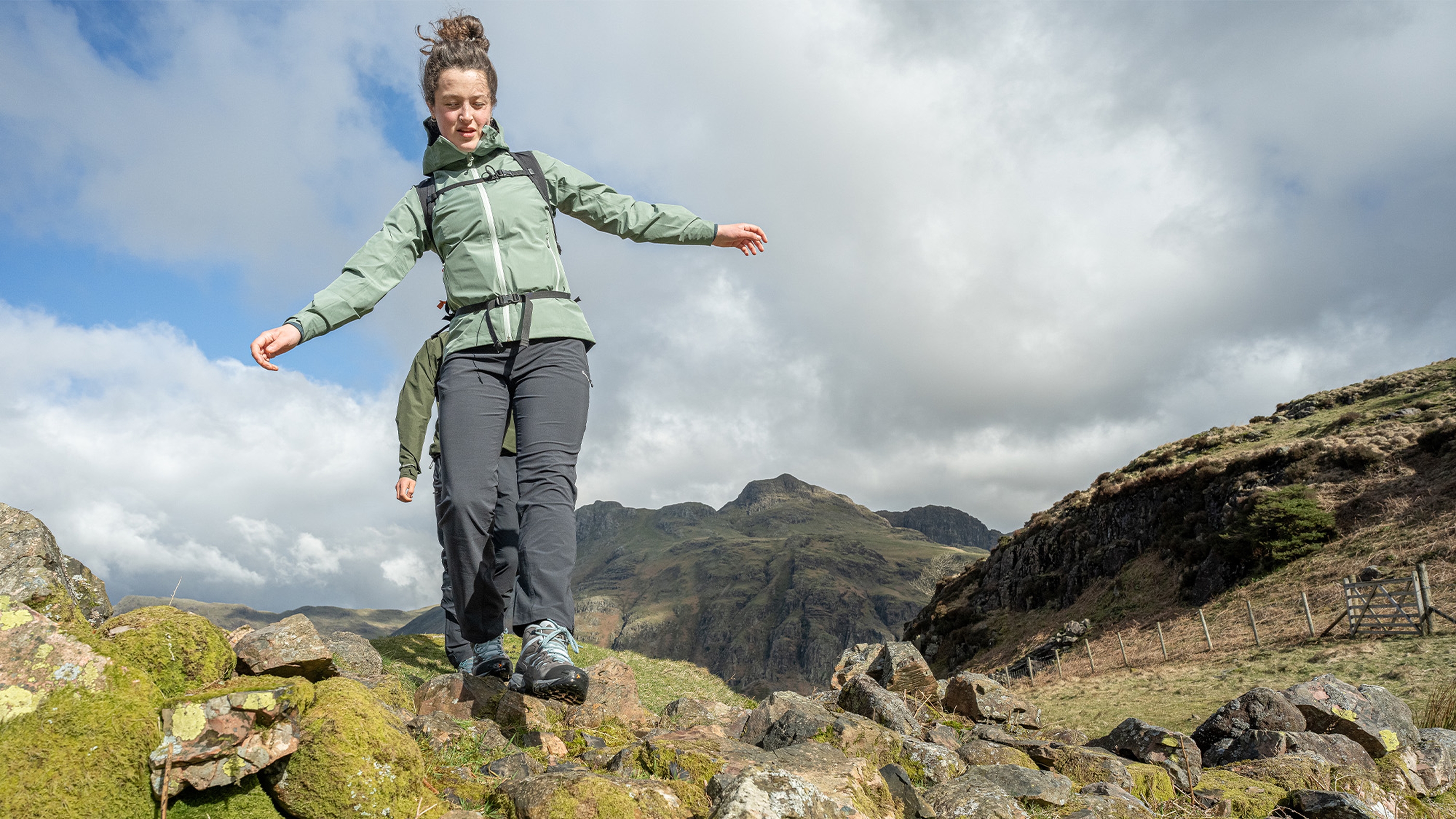 Woman hiking over rocky terrain in mountain landscape, wearing a green jacket and grey trousers, with cloudy sky in the background.
