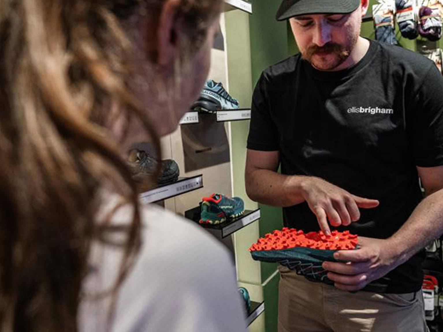A store employee shows a customer a shoe with a bright red, textured sole, surrounded by shelves of various shoes and outdoor gear.