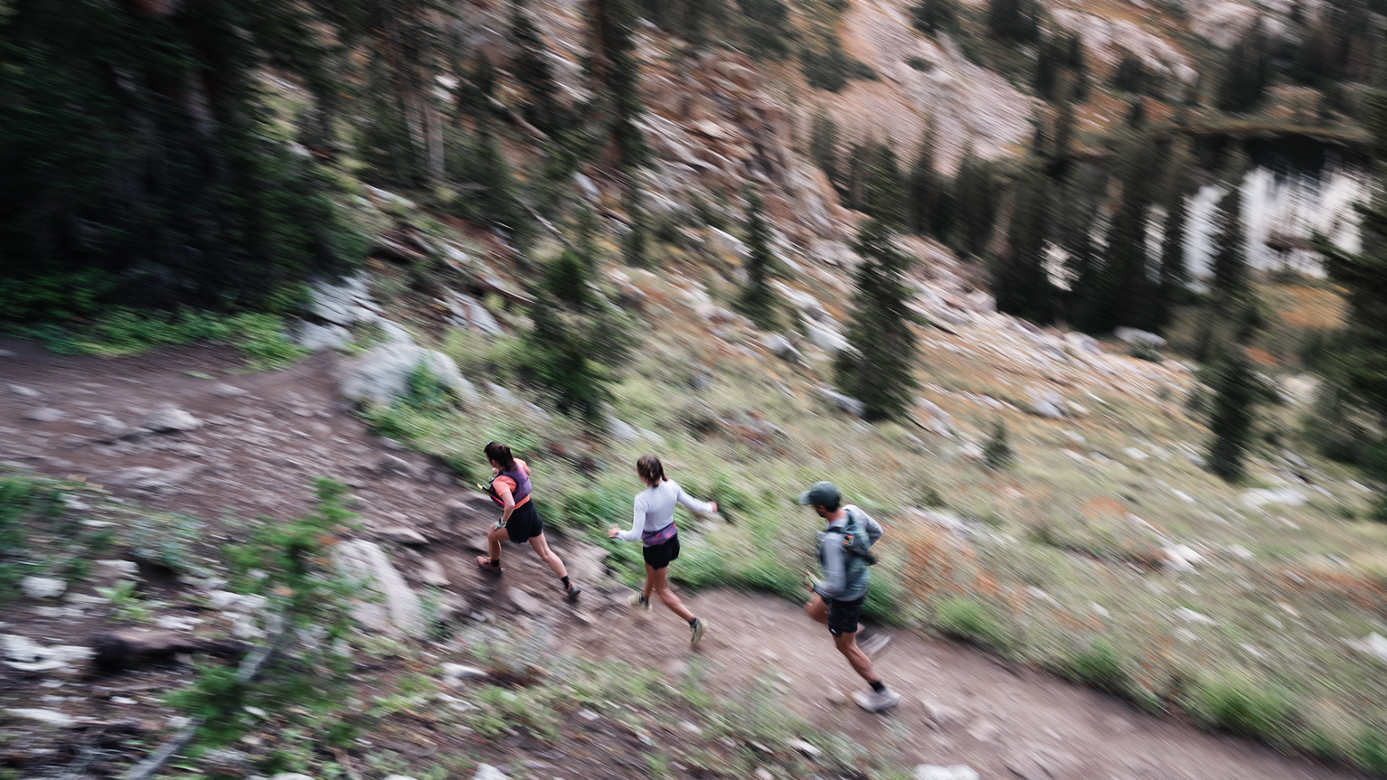 Three people trail running on a rugged mountain path, surrounded by trees and rocky terrain.