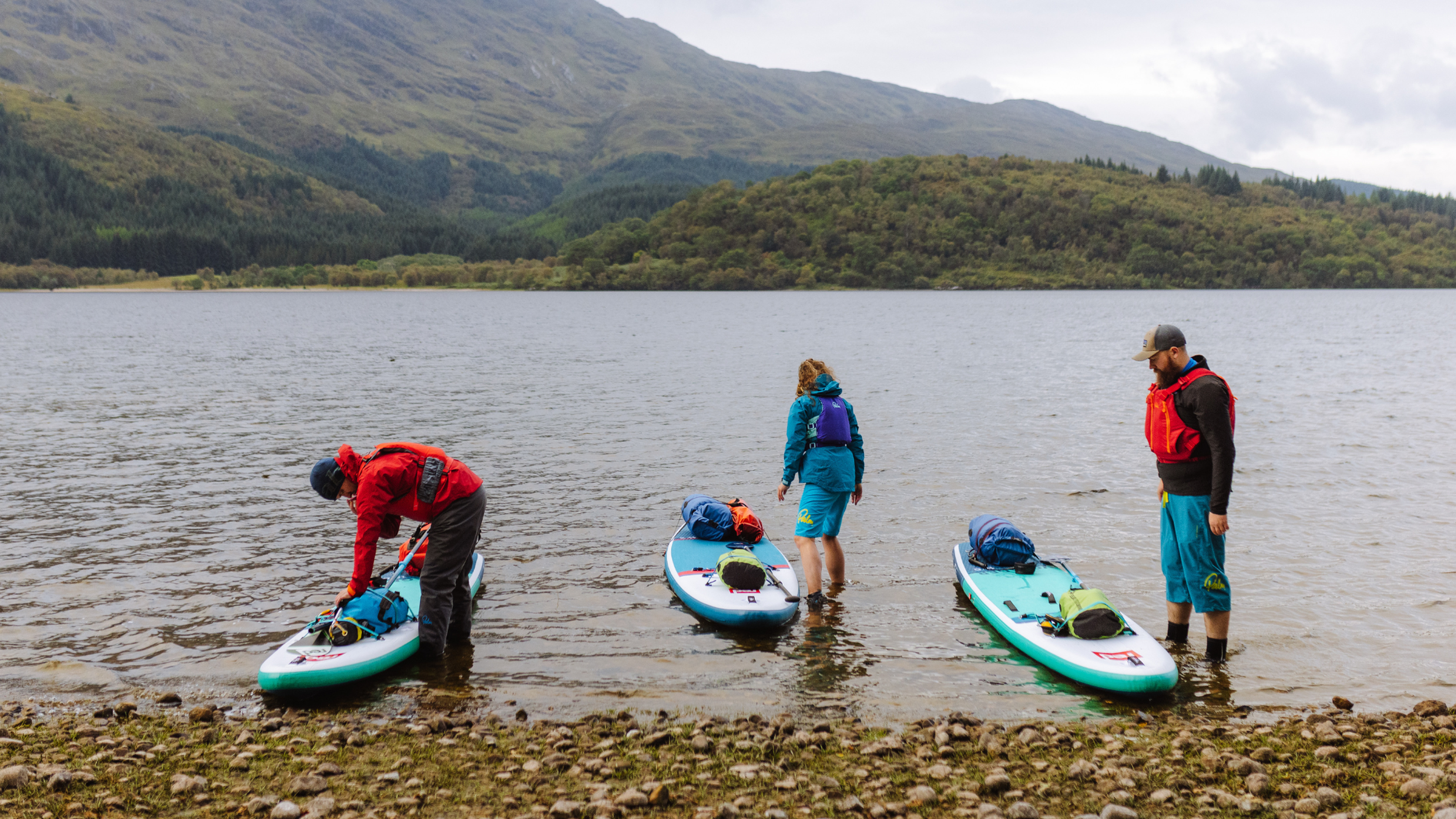 Three people in water with paddleboards near a rocky shore, surrounded by hills and a cloudy sky.