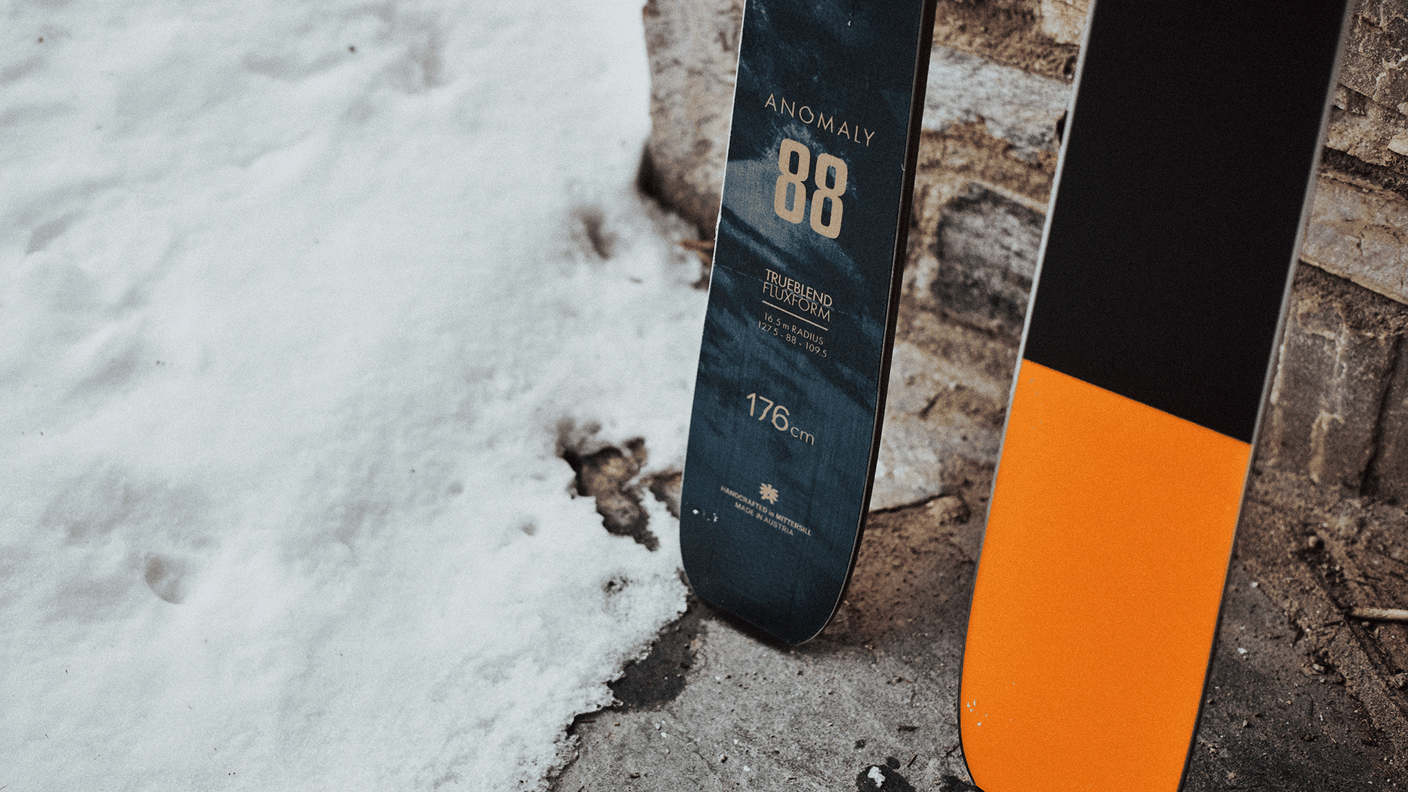 A pair of skis stand upright on snow, featuring a design with "Anomaly 88" and "176 cm" markings on the blue ski.