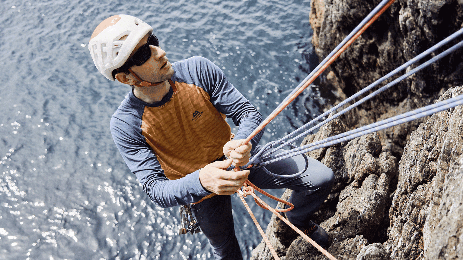 Climber in helmet and sunglasses secures ropes on a rocky cliff with the sea in the background.