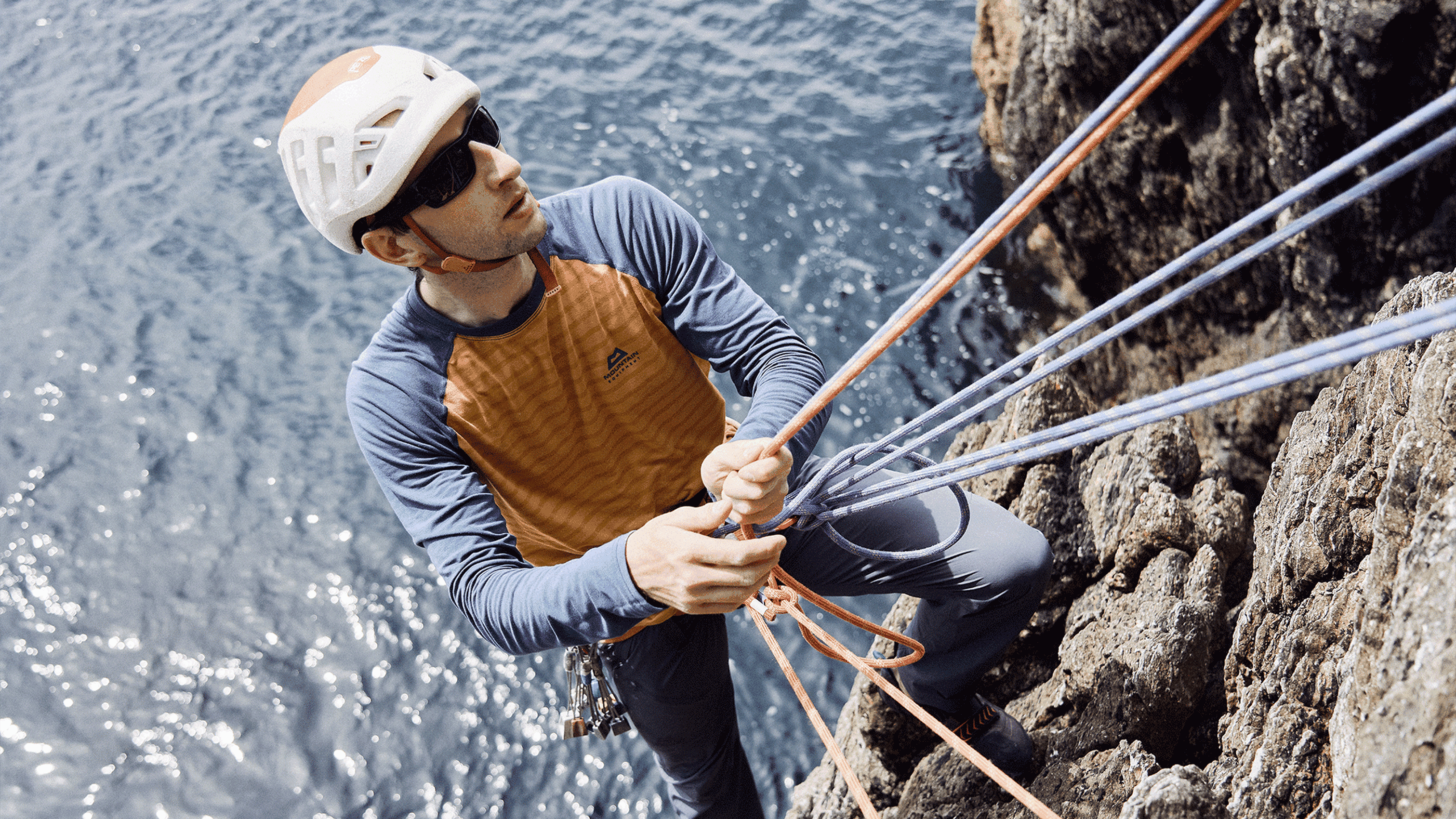 Climber in helmet and sunglasses secures ropes on a rocky cliff with the sea in the background.