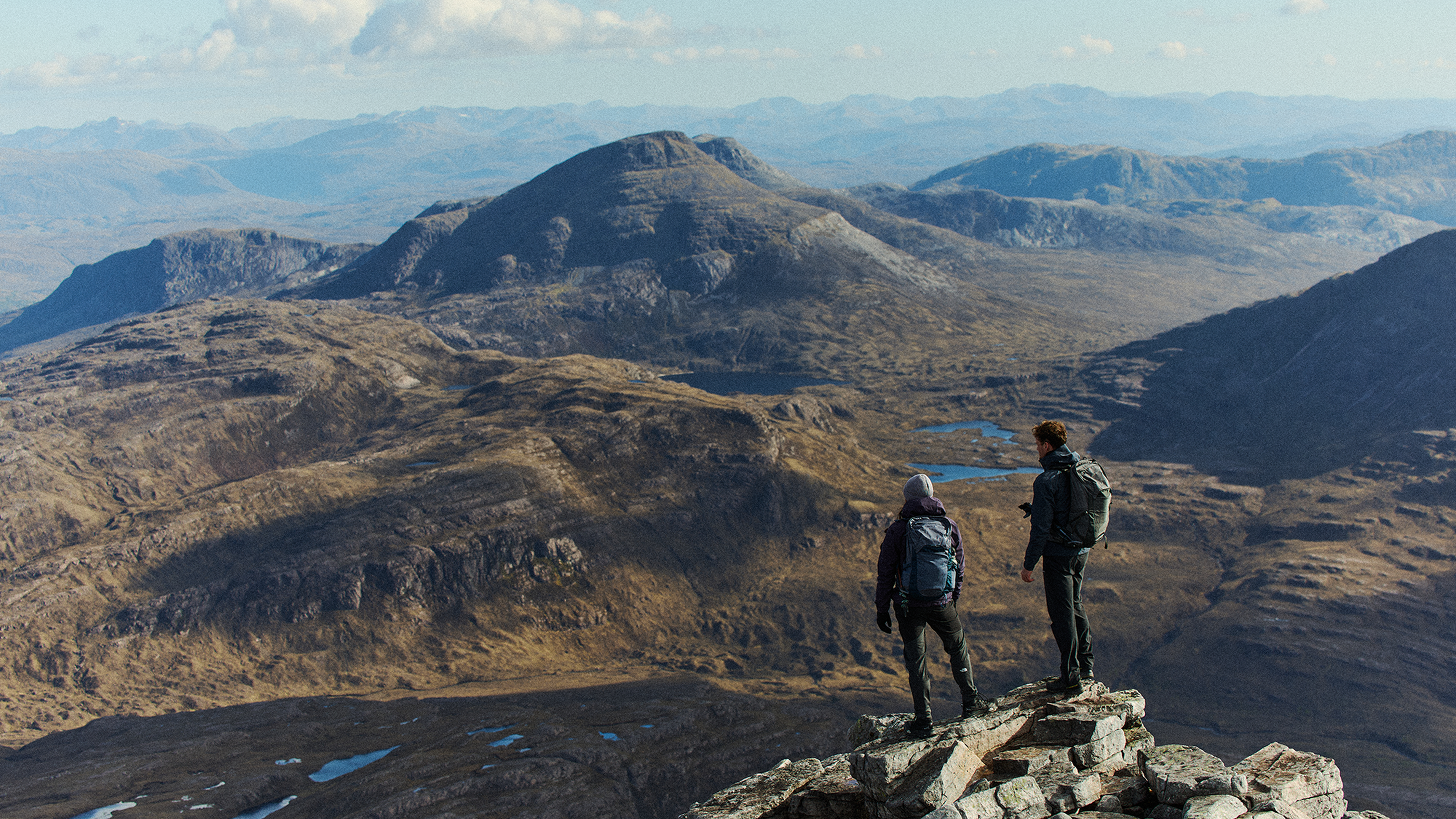 Two hikers stand on a rocky mountain peak, overlooking vast rugged terrain and distant hills under a clear blue sky.