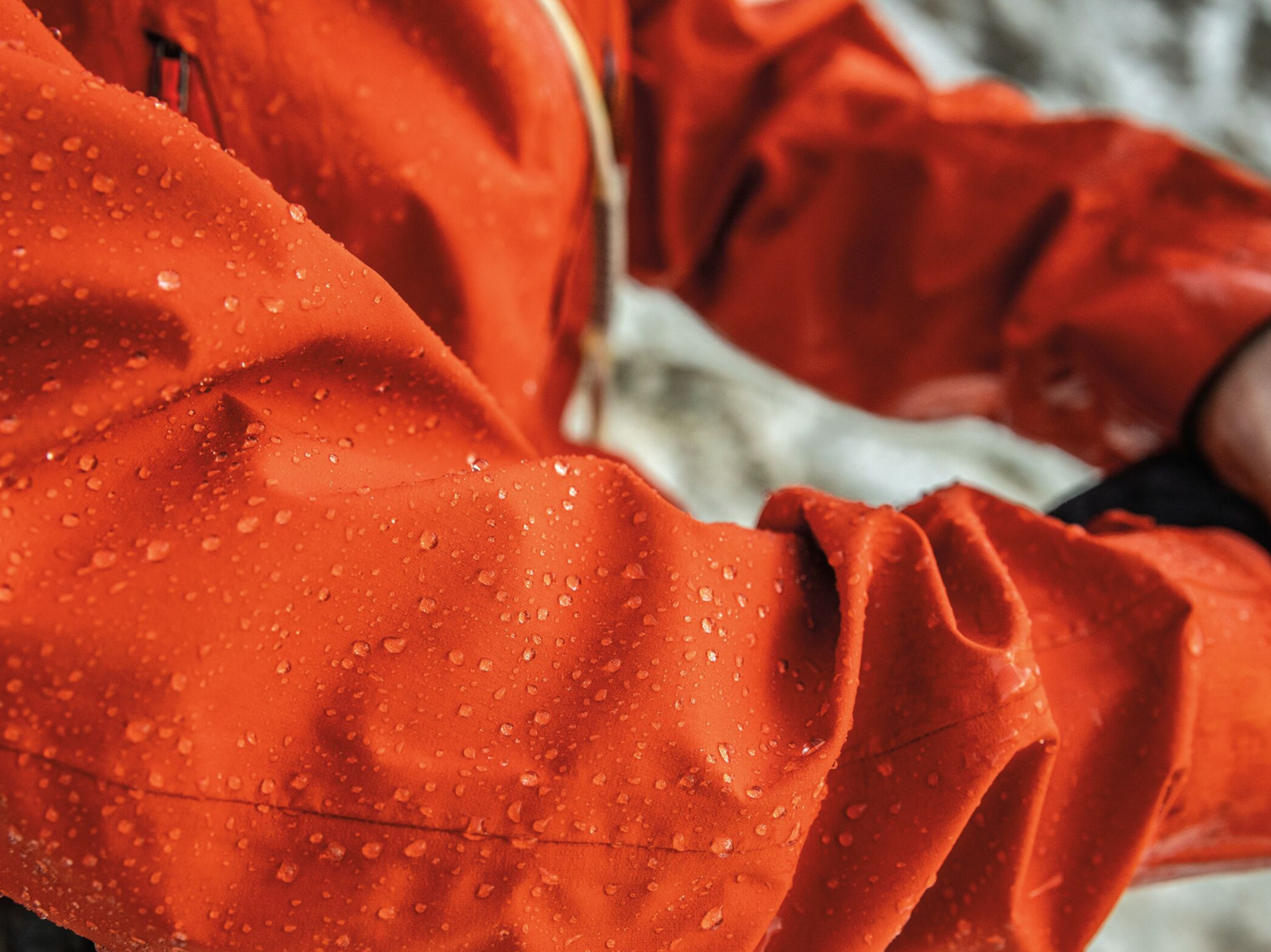 Close-up of a person adjusting the cuff of a vibrant orange waterproof jacket, with water droplets visible on the fabric.
