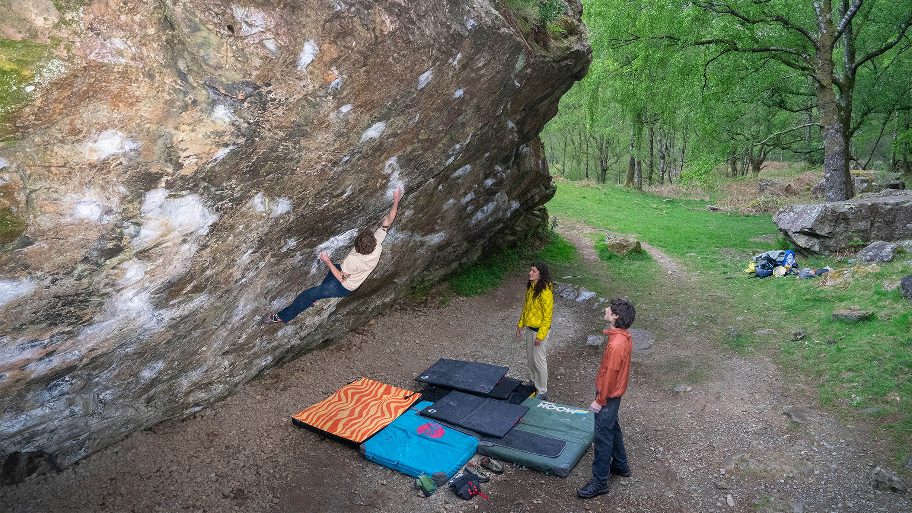 Climber bouldering on a large rock, supported by crash pads, with two people watching, set in a lush forest area.