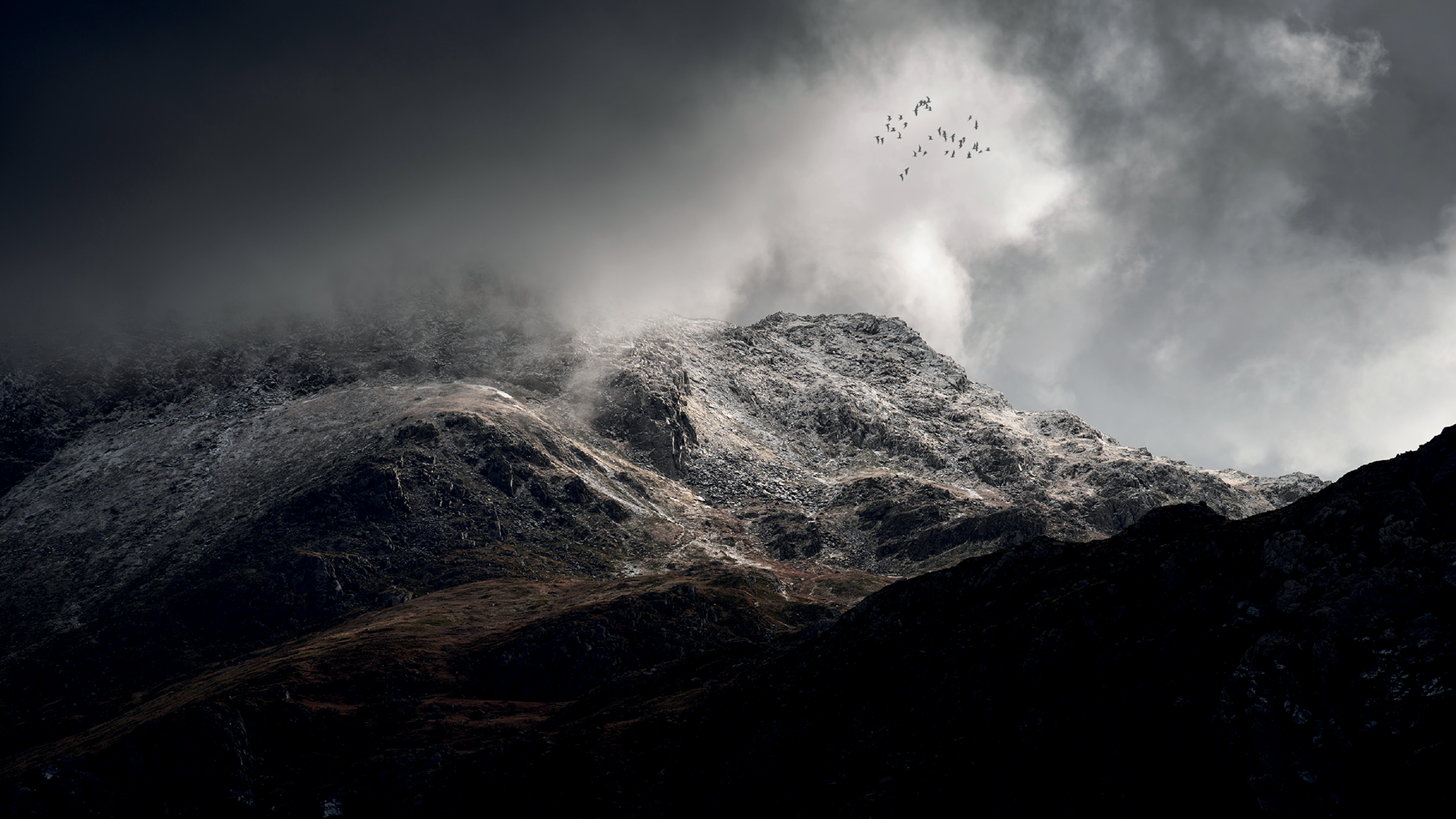 A rugged mountain peak partially covered in mist, with a flock of birds flying overhead against a dramatic cloudy sky.